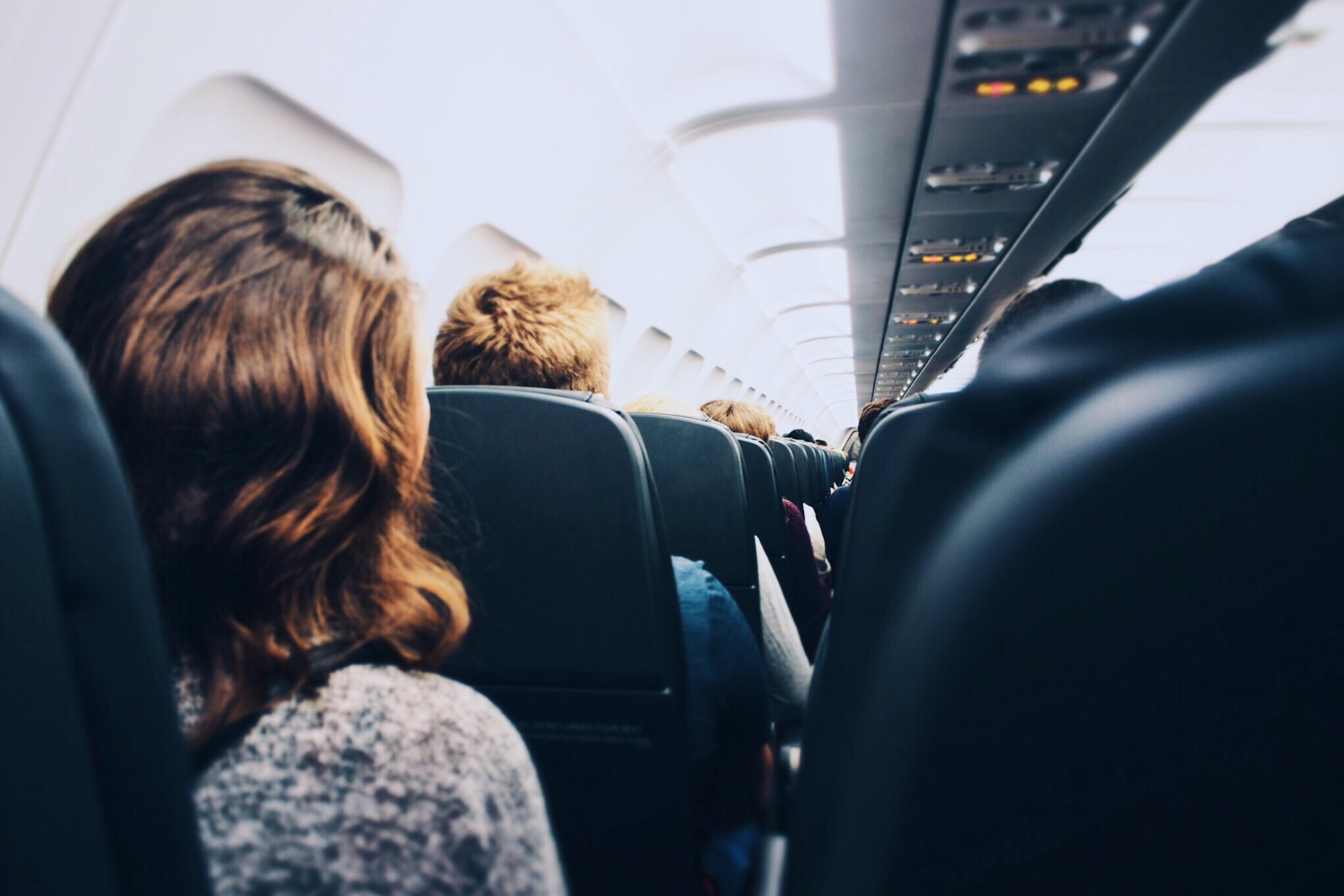 Photo of a plane cabin interior showing passengers seated, viewed from the back.