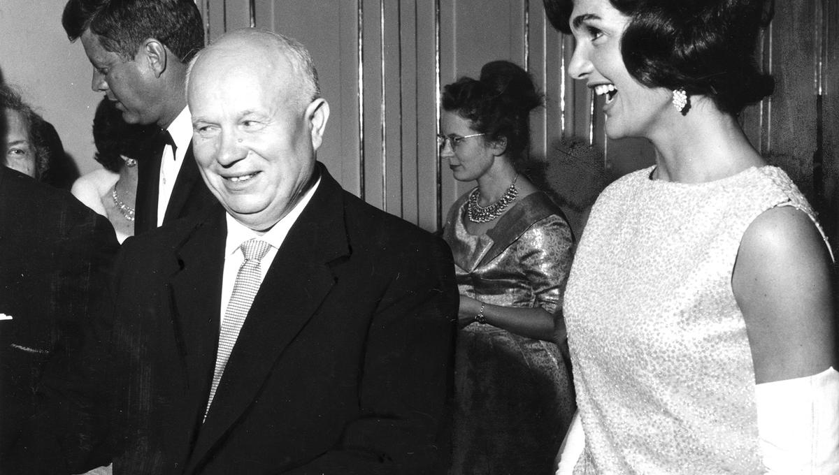 A black-and-white photo of a man in a suit and tie smiling while a woman in a glittery dress and gloves laughs beside him. A man in a tuxedo and a woman in a formal dress and glasses are visible in the background at a social gathering.