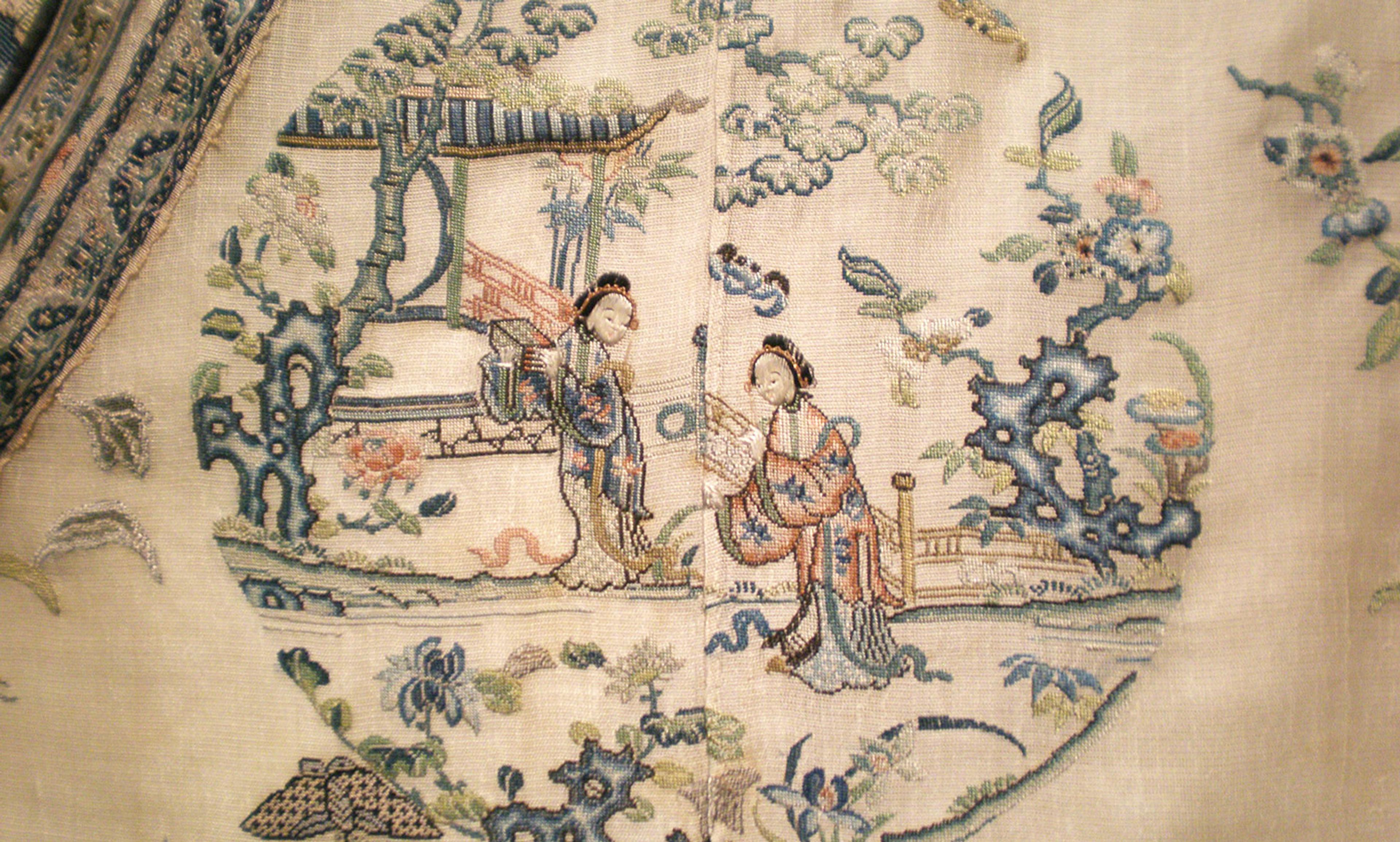 Traditional Chinese embroidery depicting two figures in a garden with trees and a pavilion on a beige fabric.