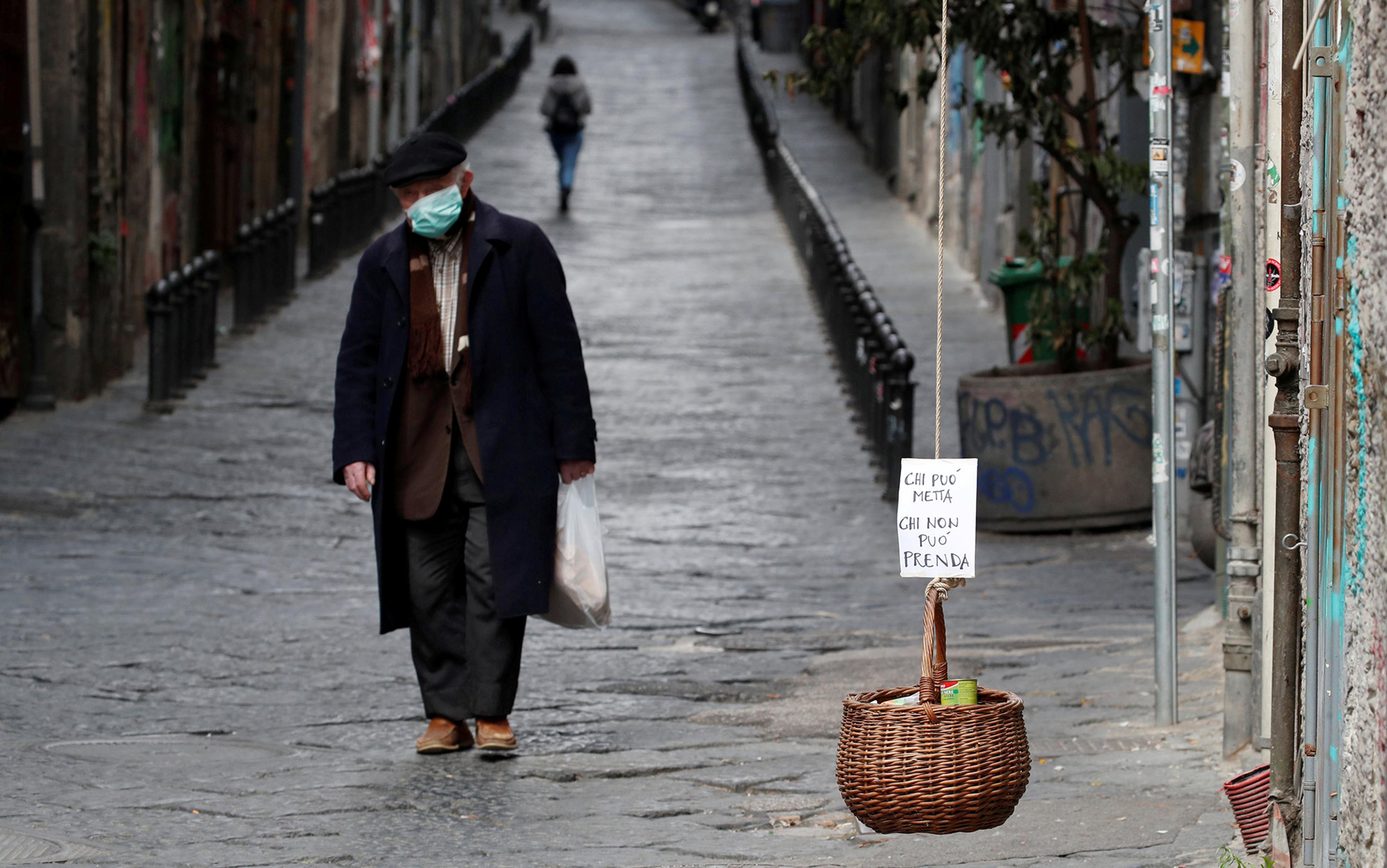 An elderly man wearing a face mask walking down a cobbled street with a hanging wicker basket for donations in the foreground.