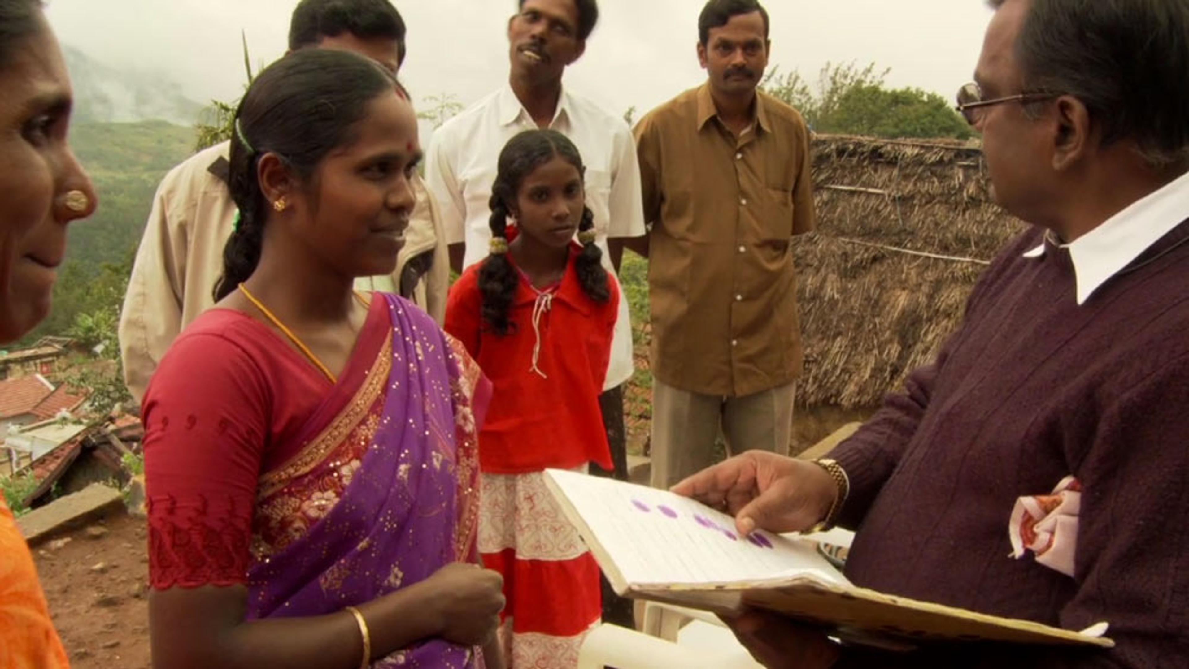 A group outdoors, a woman in a purple sari interacts with a man holding a document, with others watching in the background.