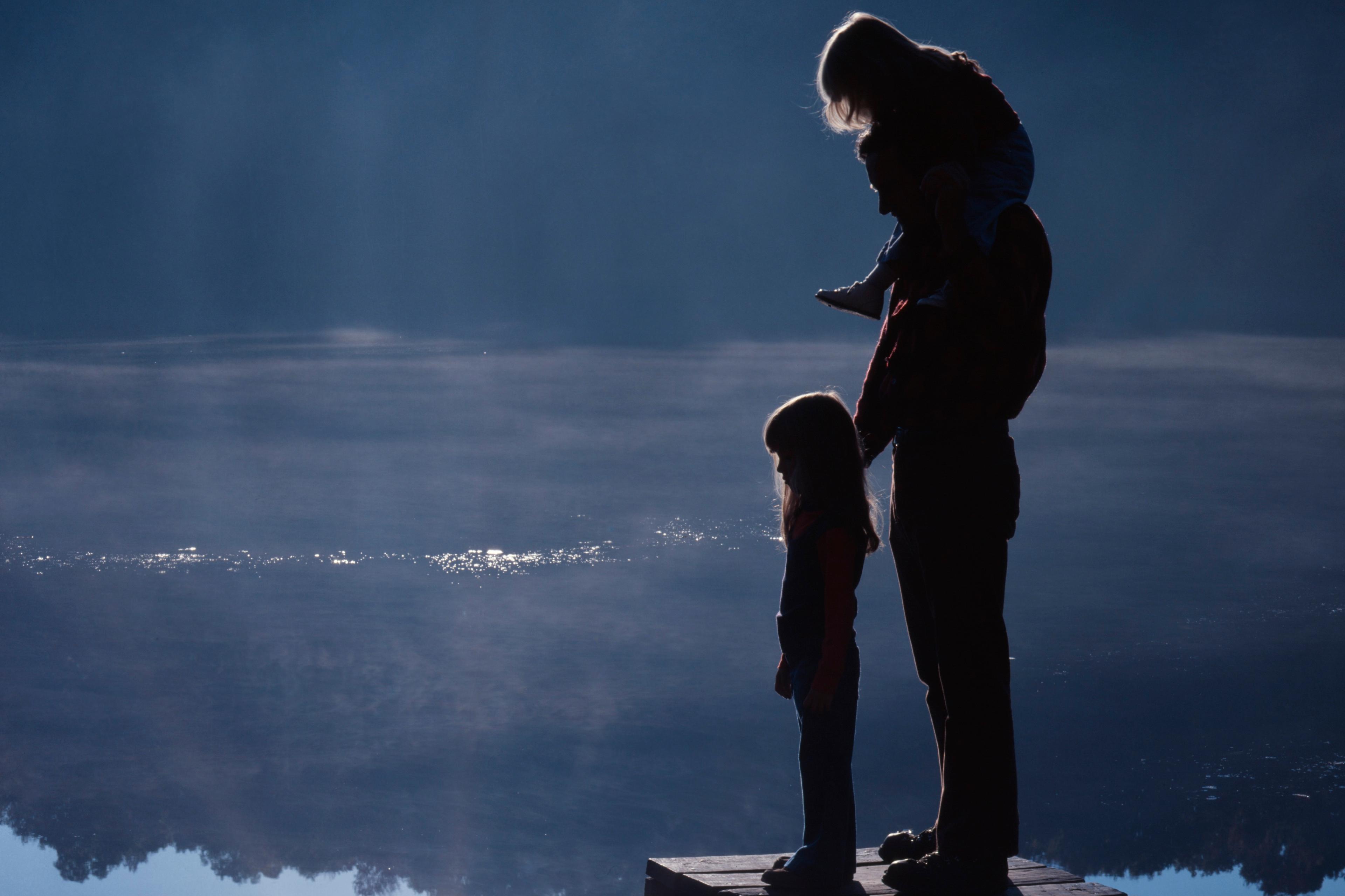 Silhouetted photo of a family on a wooden pier overlooking a serene lake at sunset.