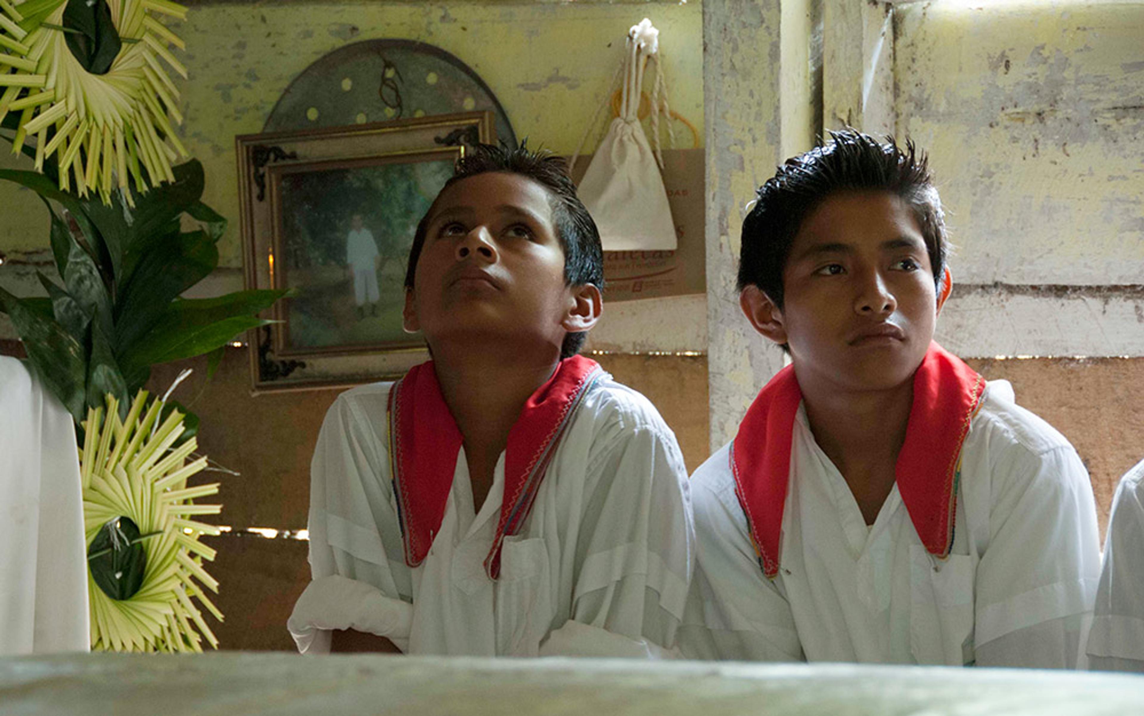 Two boys in white shirts with red collars sitting indoors looking thoughtful with a framed picture on the wall behind them.