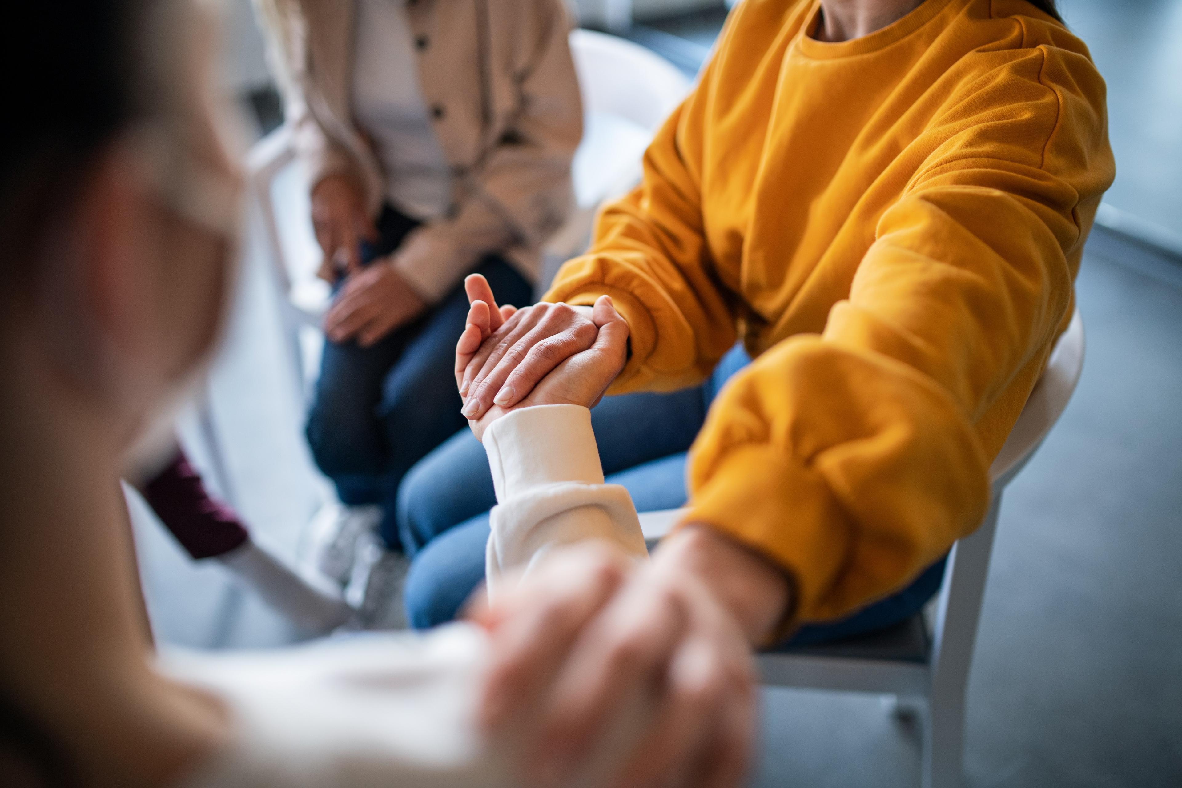 Photo of people sitting in a group holding hands, one wearing a yellow jumper, conveying support and connection.