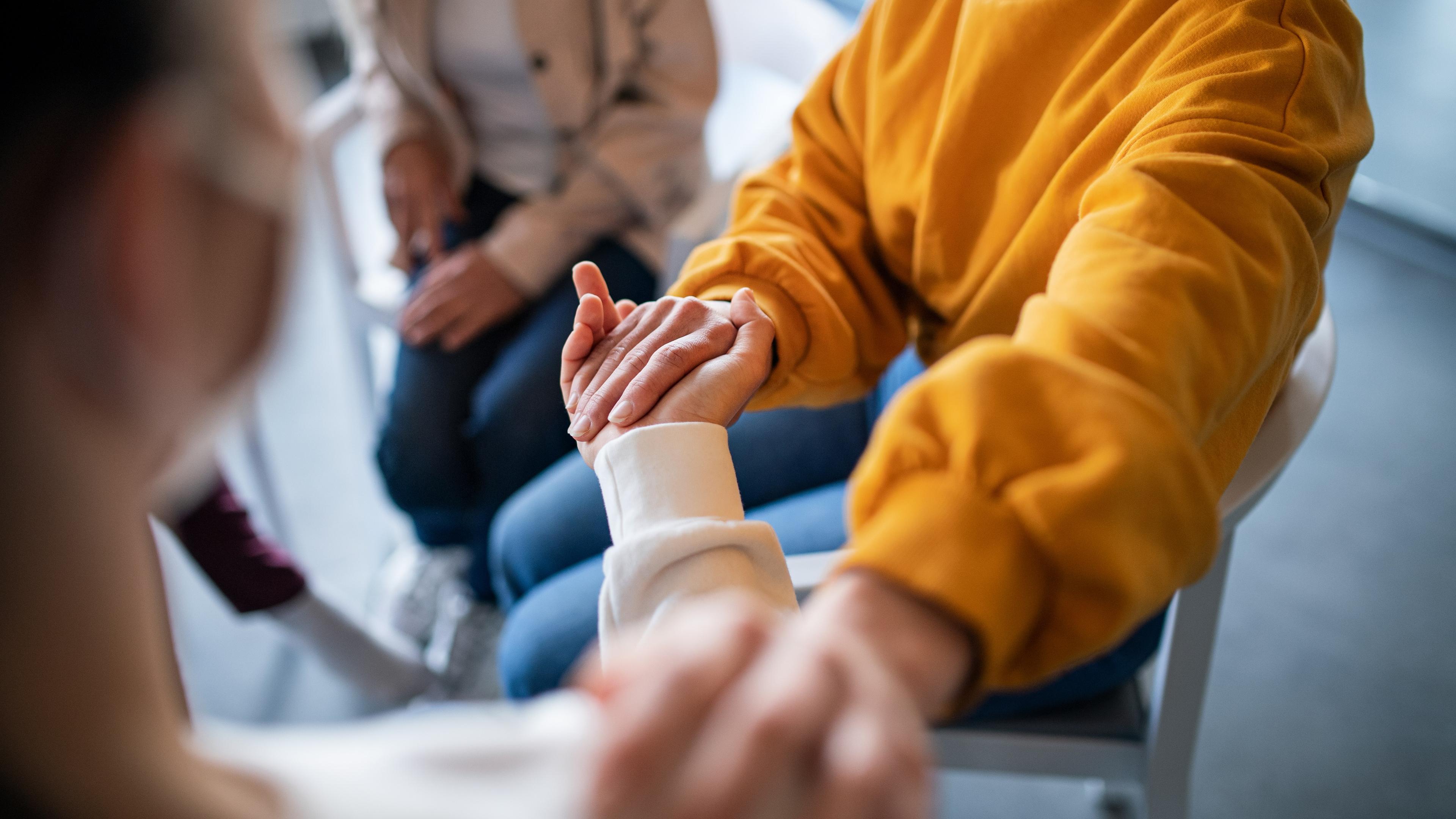 Photo of people sitting in a group holding hands, one wearing a yellow jumper, conveying support and connection.