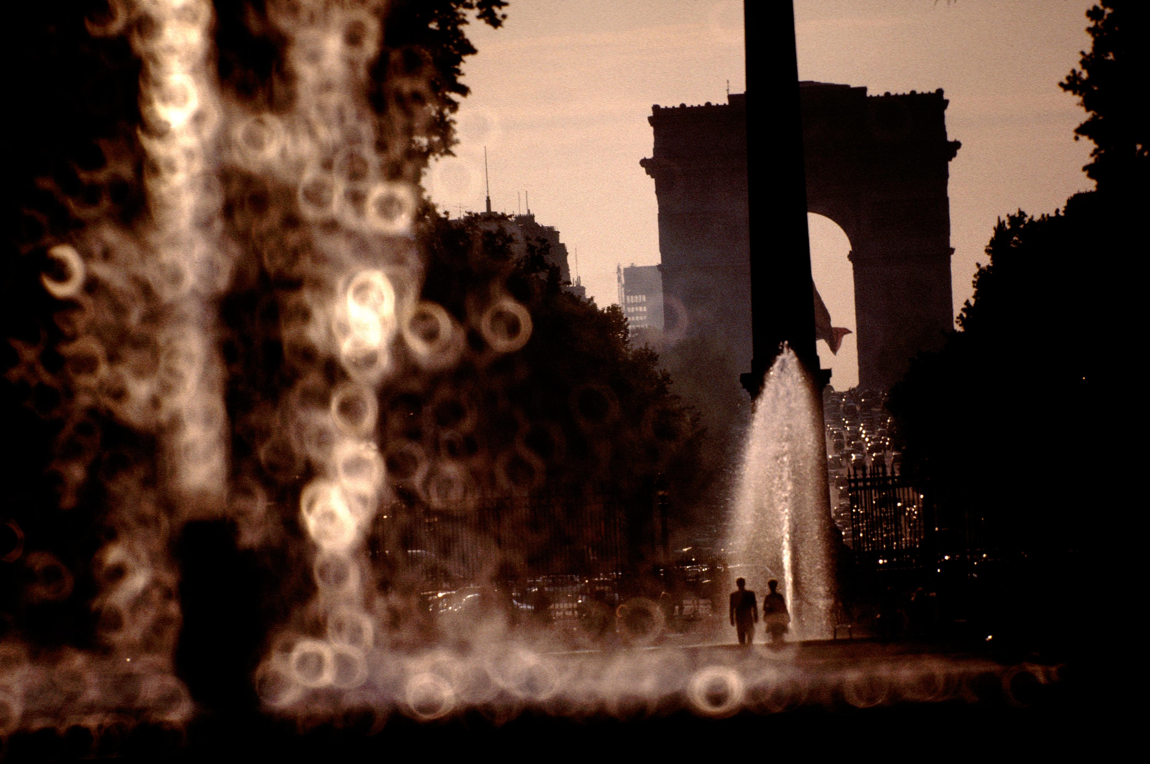Photo of two silhouettes walking by fountains with bokeh effect in the foreground, Arc de Triomphe in the background.