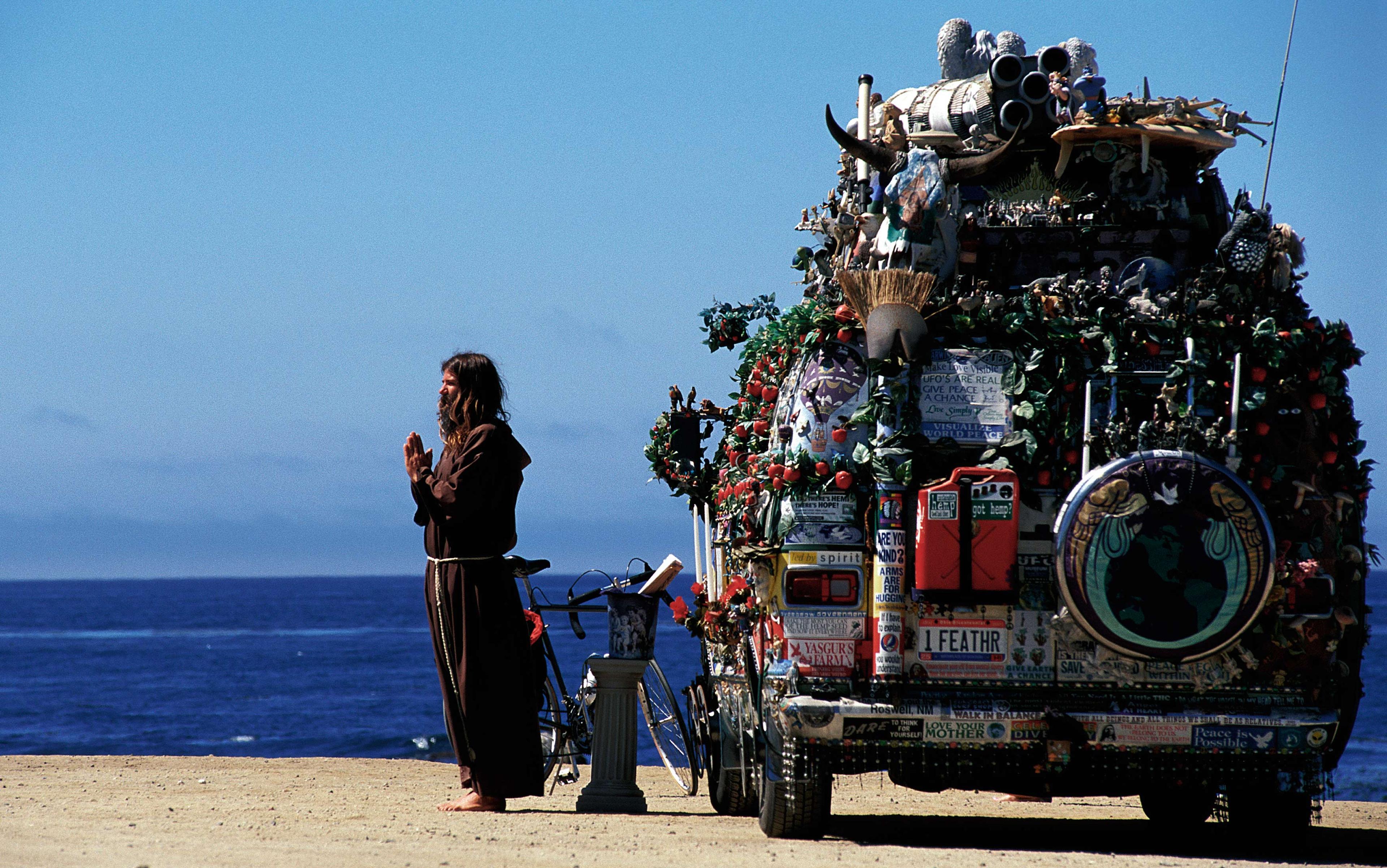 A long-haired man in brown robes standing beside a decorated vehicle by a beach, hands in prayer, with the ocean and blue sky in the background.