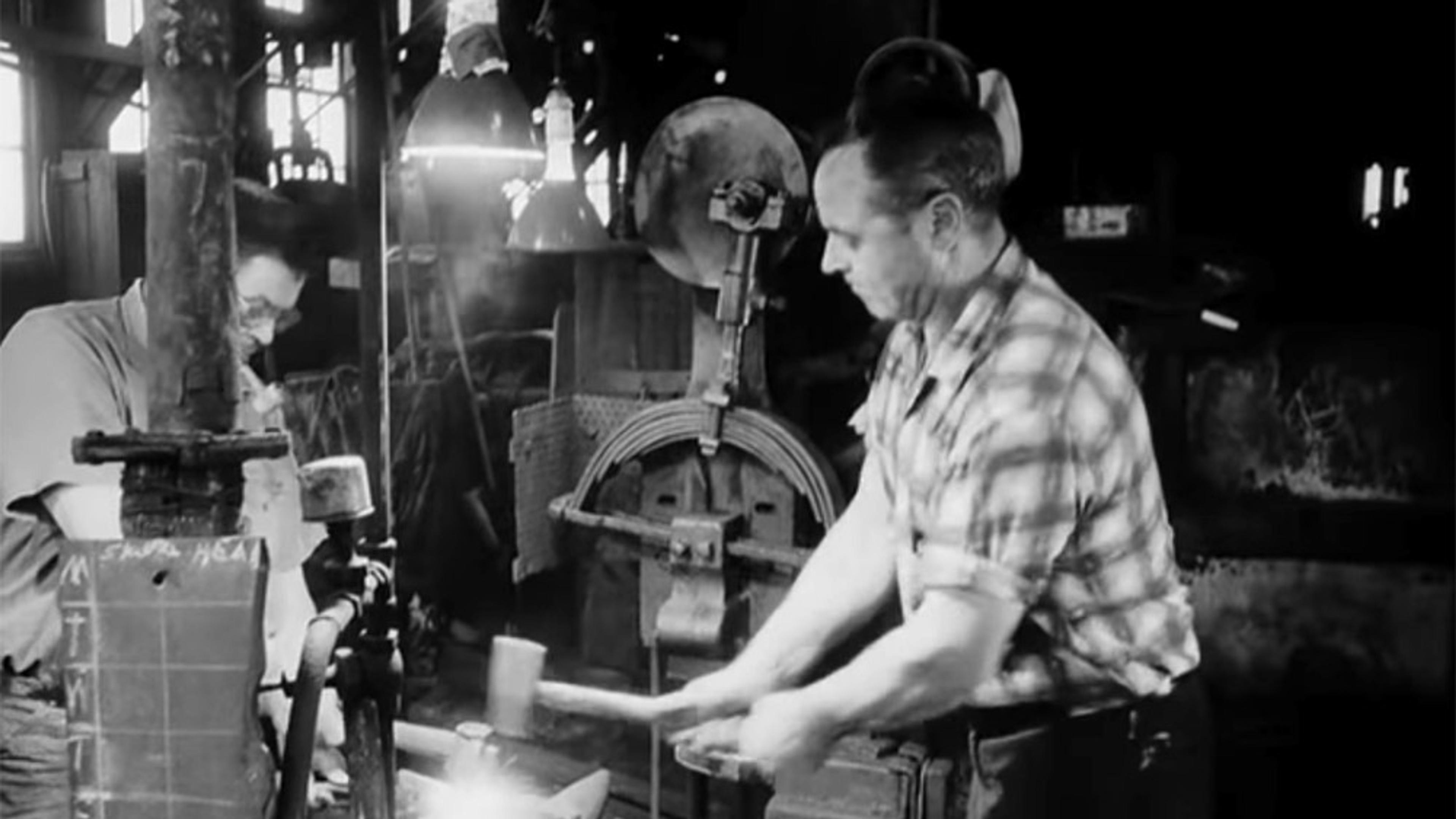 Vintage photo of two men in a workshop, one hammering a piece of metal on an anvil, the other monitoring nearby equipment.