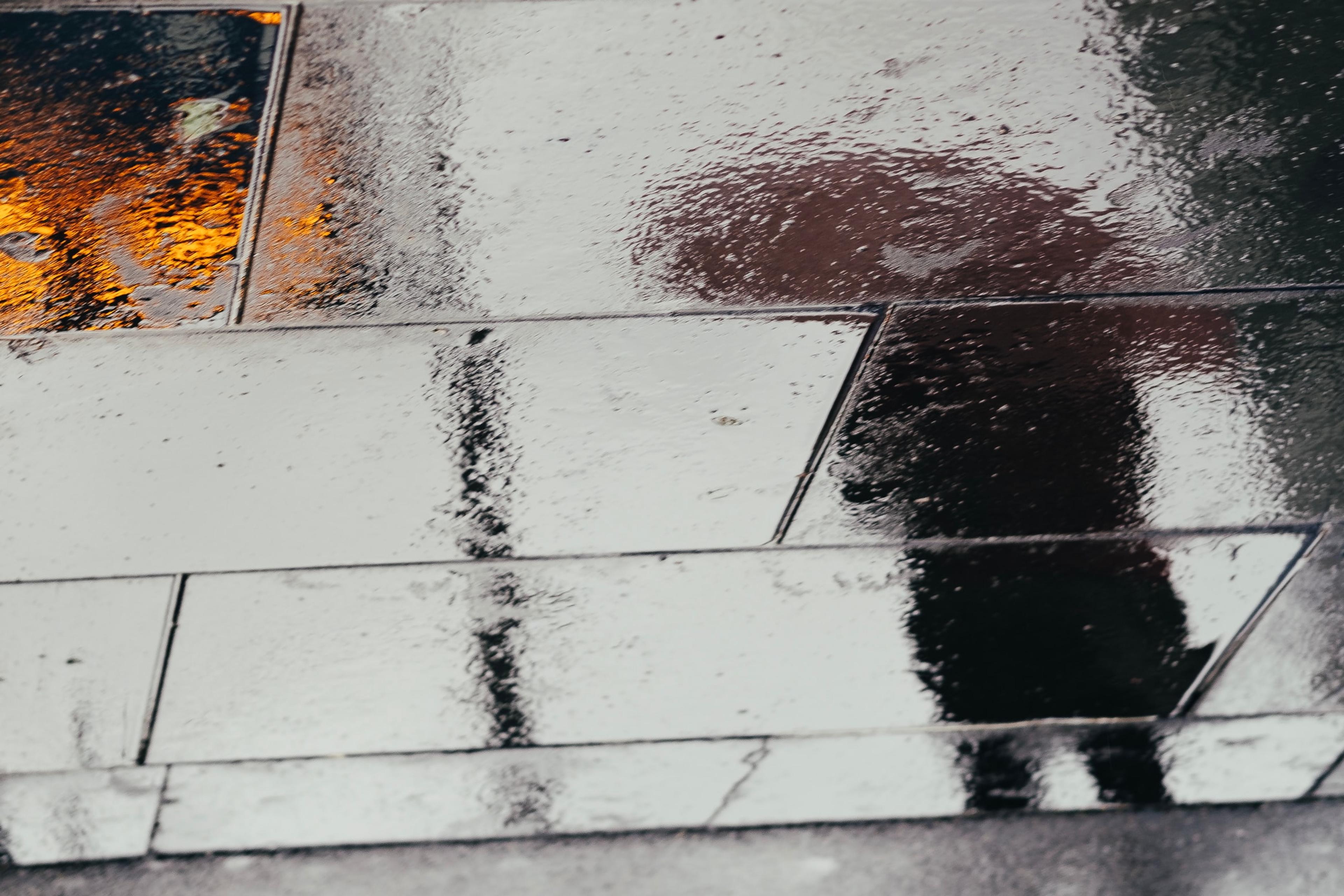 Photo of a wet pavement reflecting a person’s silhouette and orange lights on a rainy day.