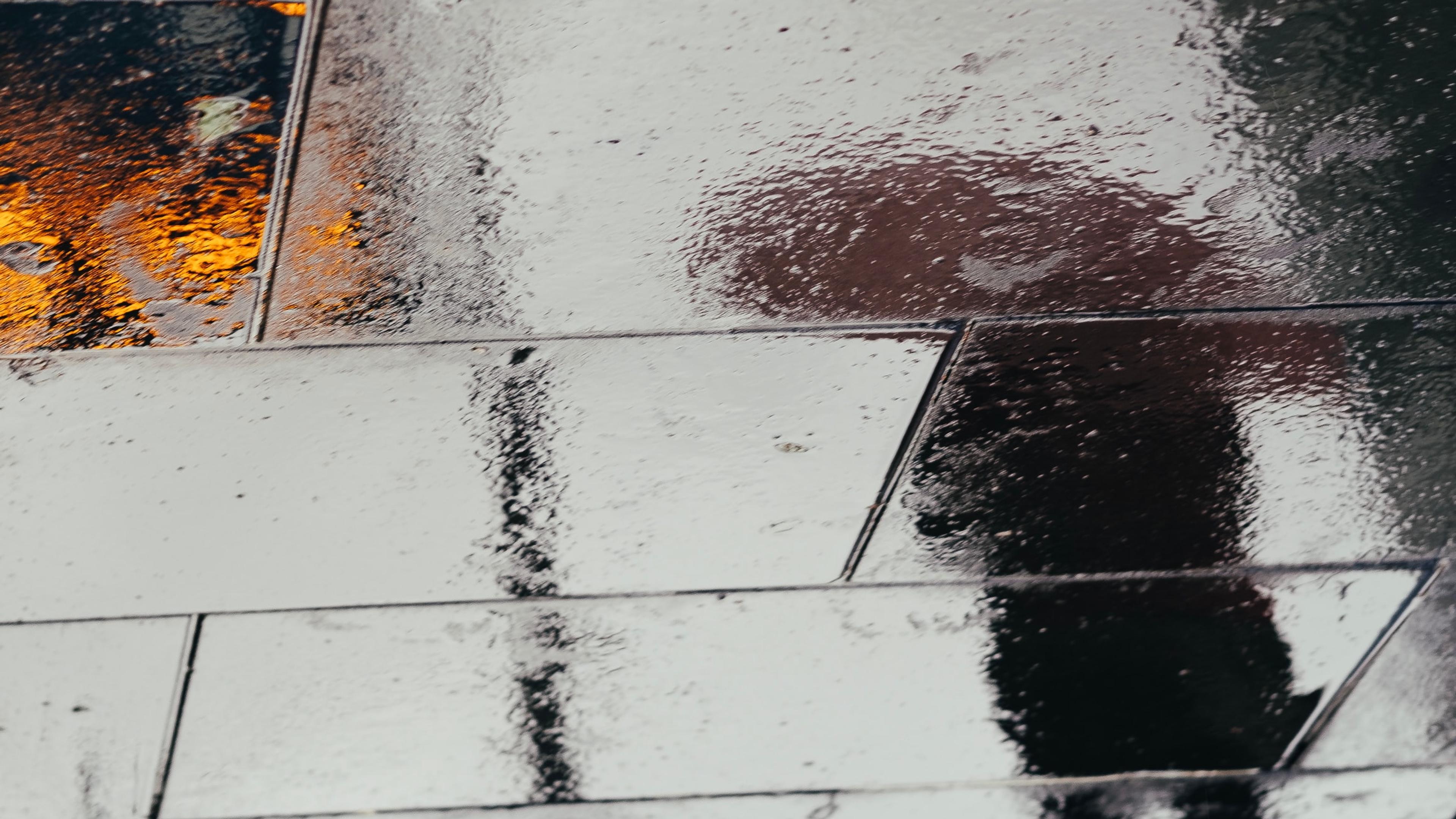 Photo of a wet pavement reflecting a person’s silhouette and orange lights on a rainy day.
