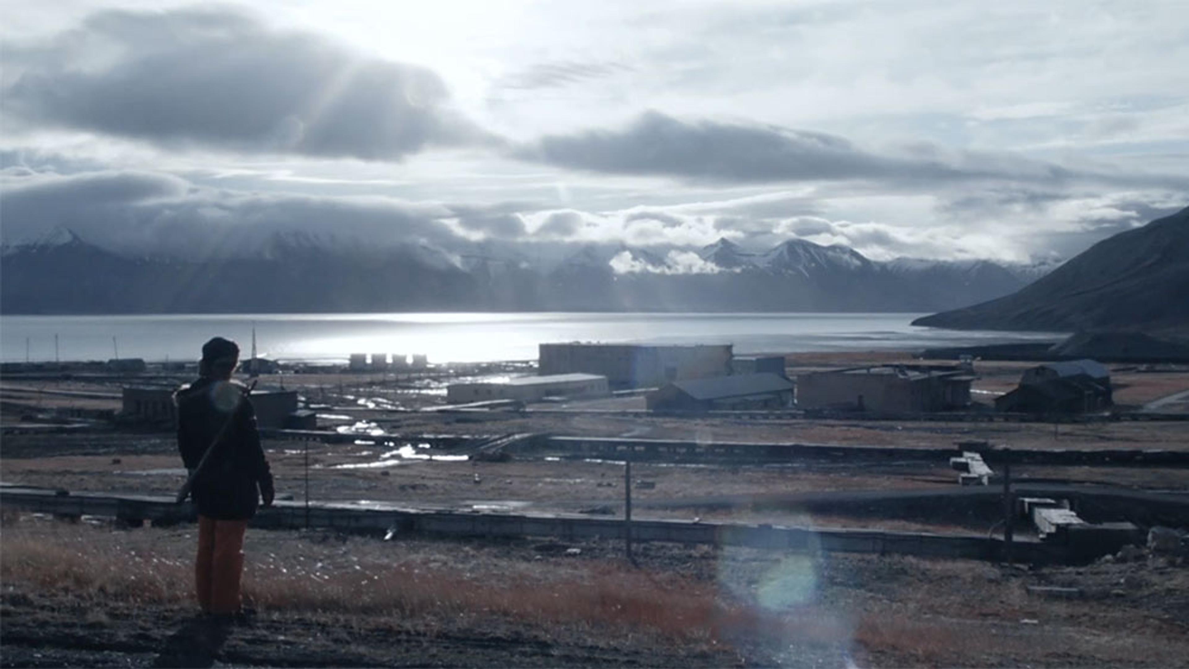 A person standing on a hill overlooking a small settlement by a lake, with snow-capped mountains under a cloudy sky in the background.