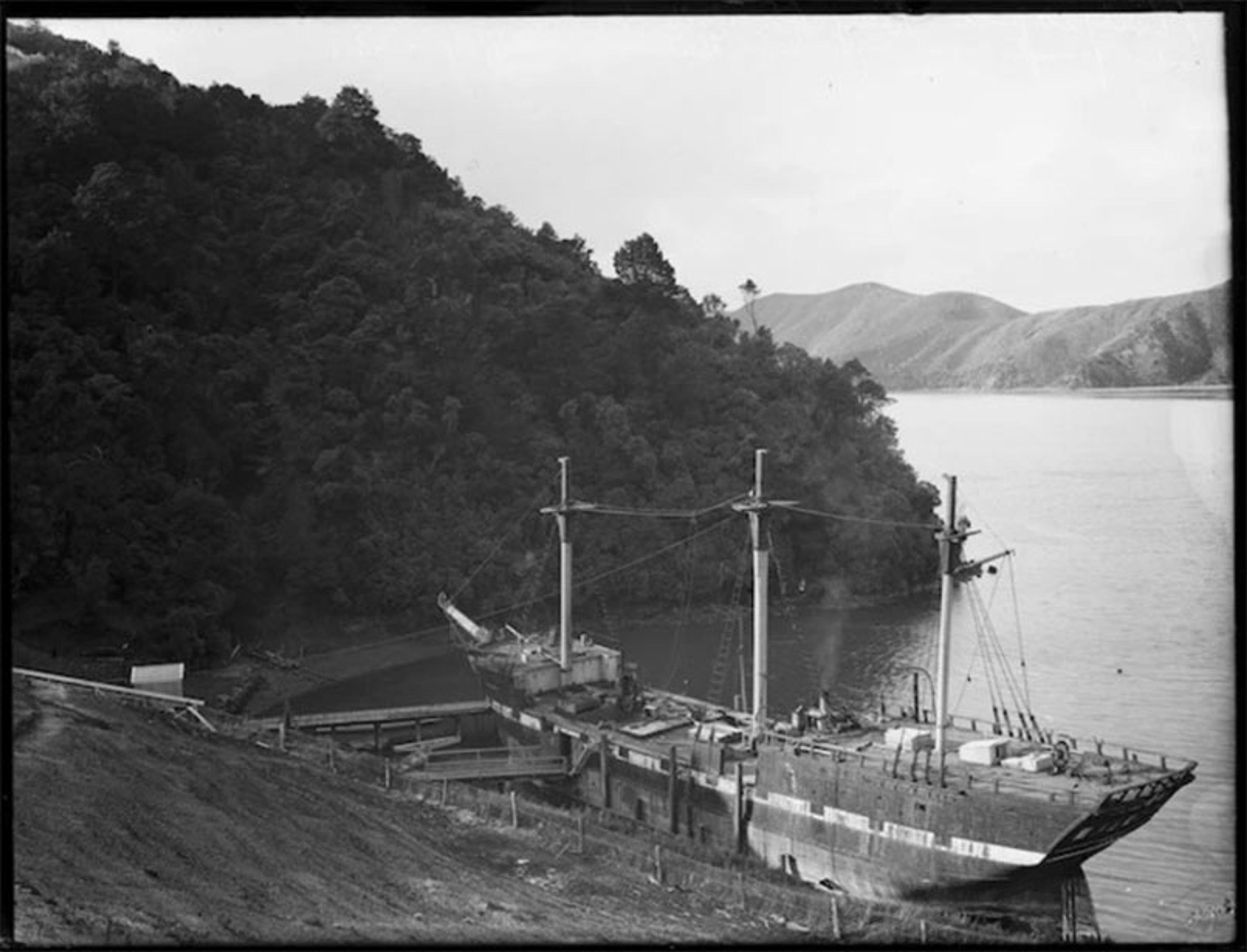 Black and white photo of an old ship docked by a wooded hillside with mountains and a river in the background.