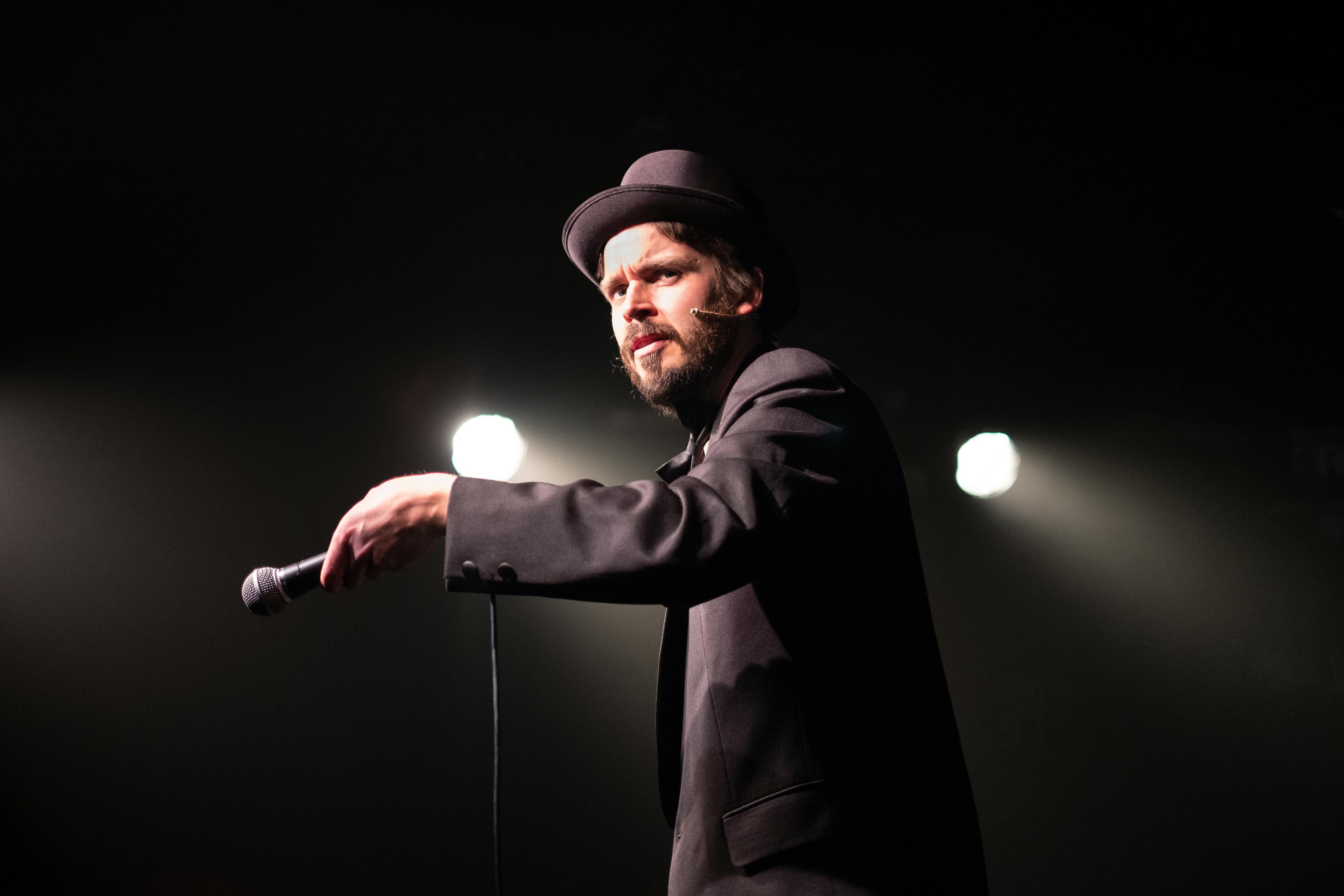 Photo of a man in a suit and bowler hat holding a microphone performing on stage with spotlights in the background.