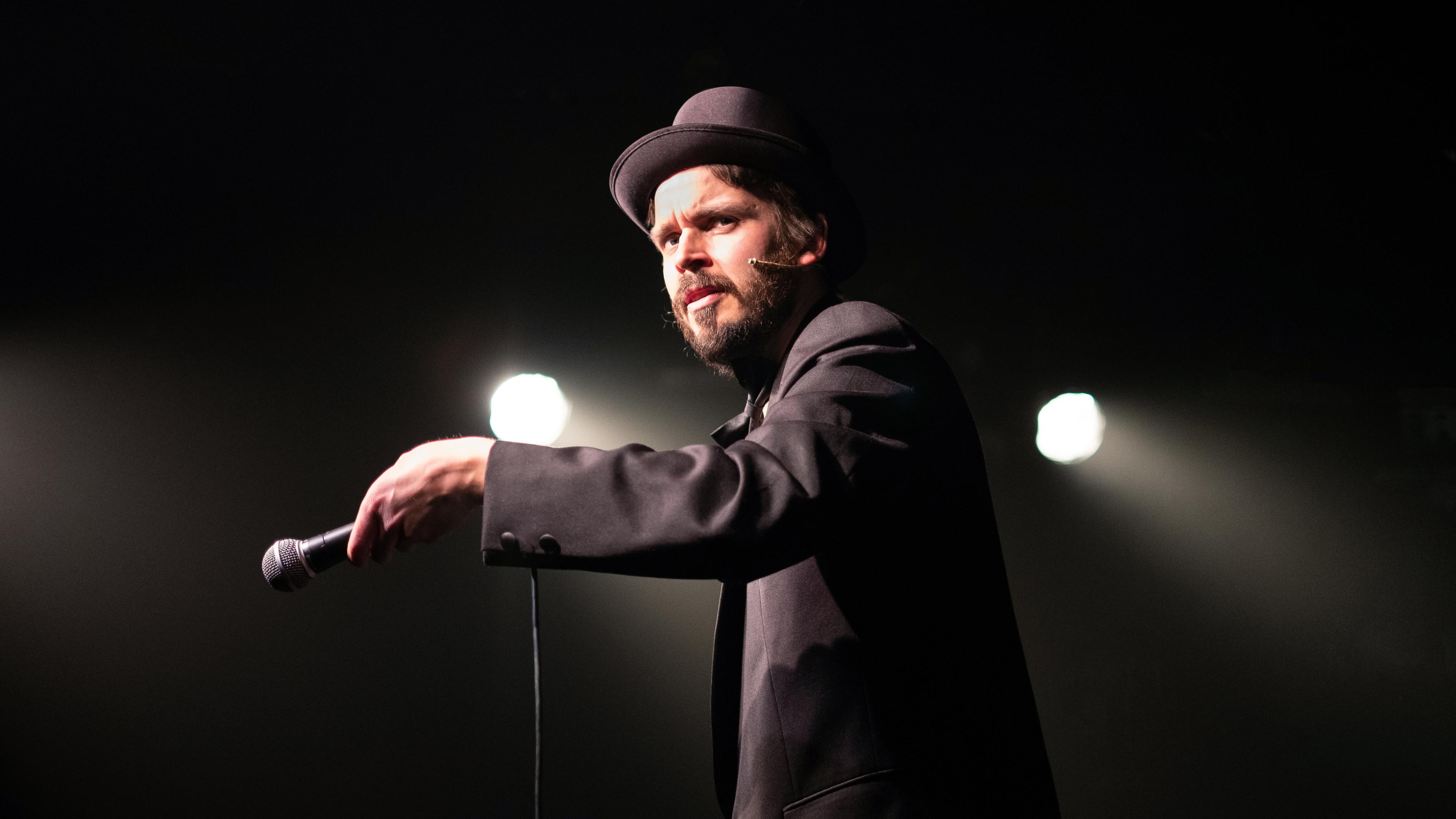 Photo of a man in a suit and bowler hat holding a microphone performing on stage with spotlights in the background.