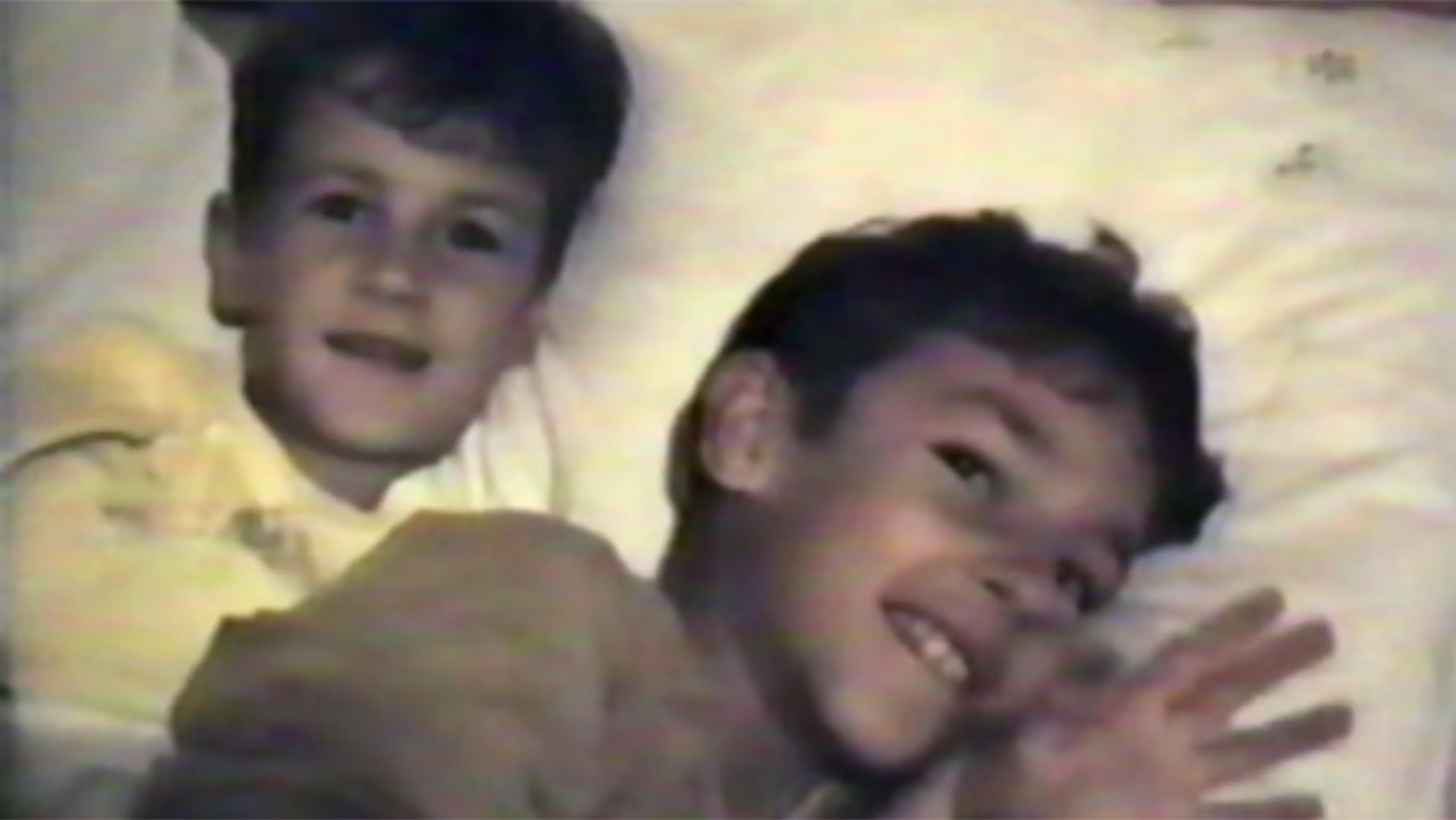 A vintage photo of two boys smiling while lying on a bed, with pillows in the background. The image has a nostalgic feel.