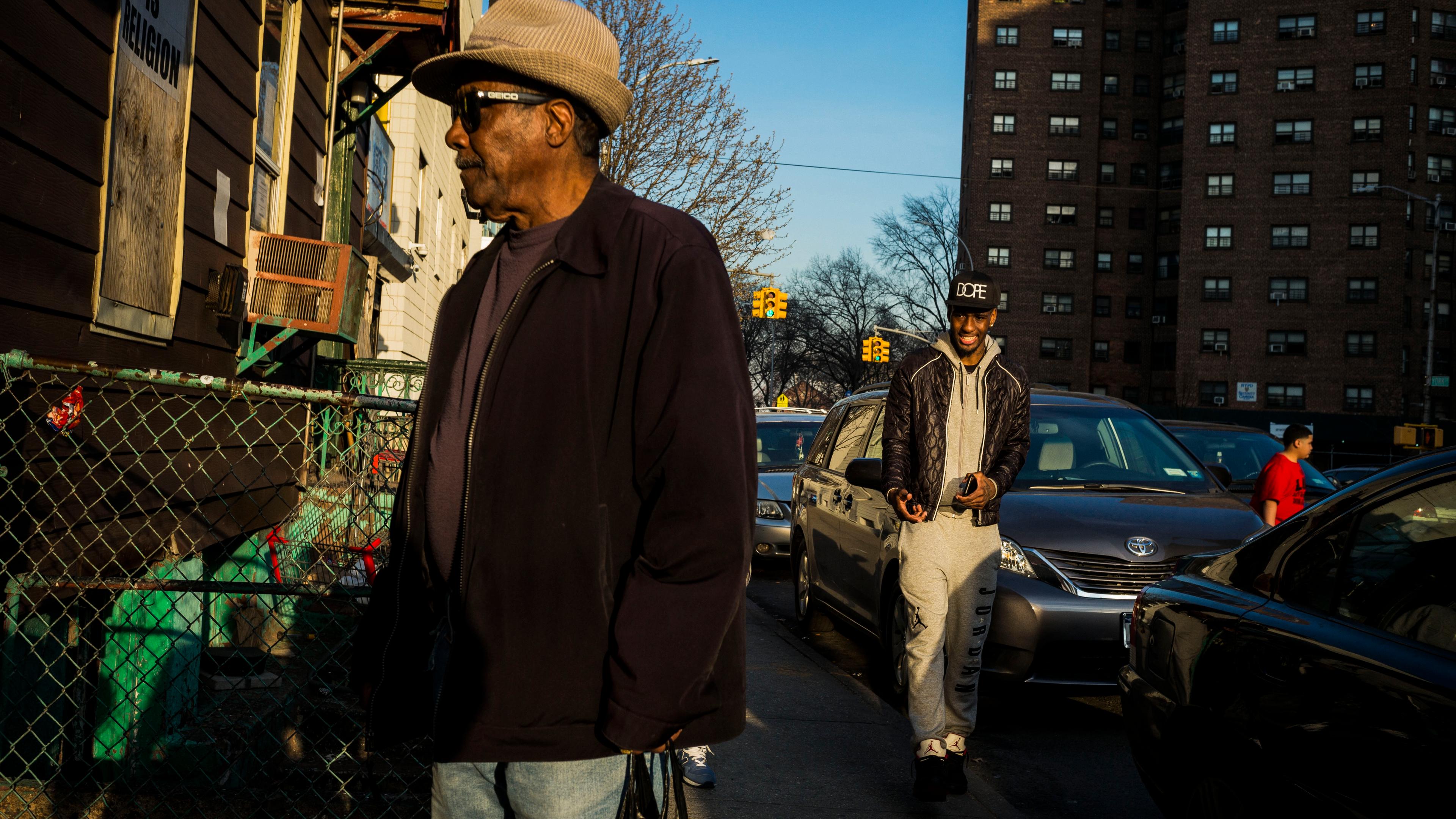 Photo of a city street with a man in a hat in the foreground and another man walking among cars in the background.