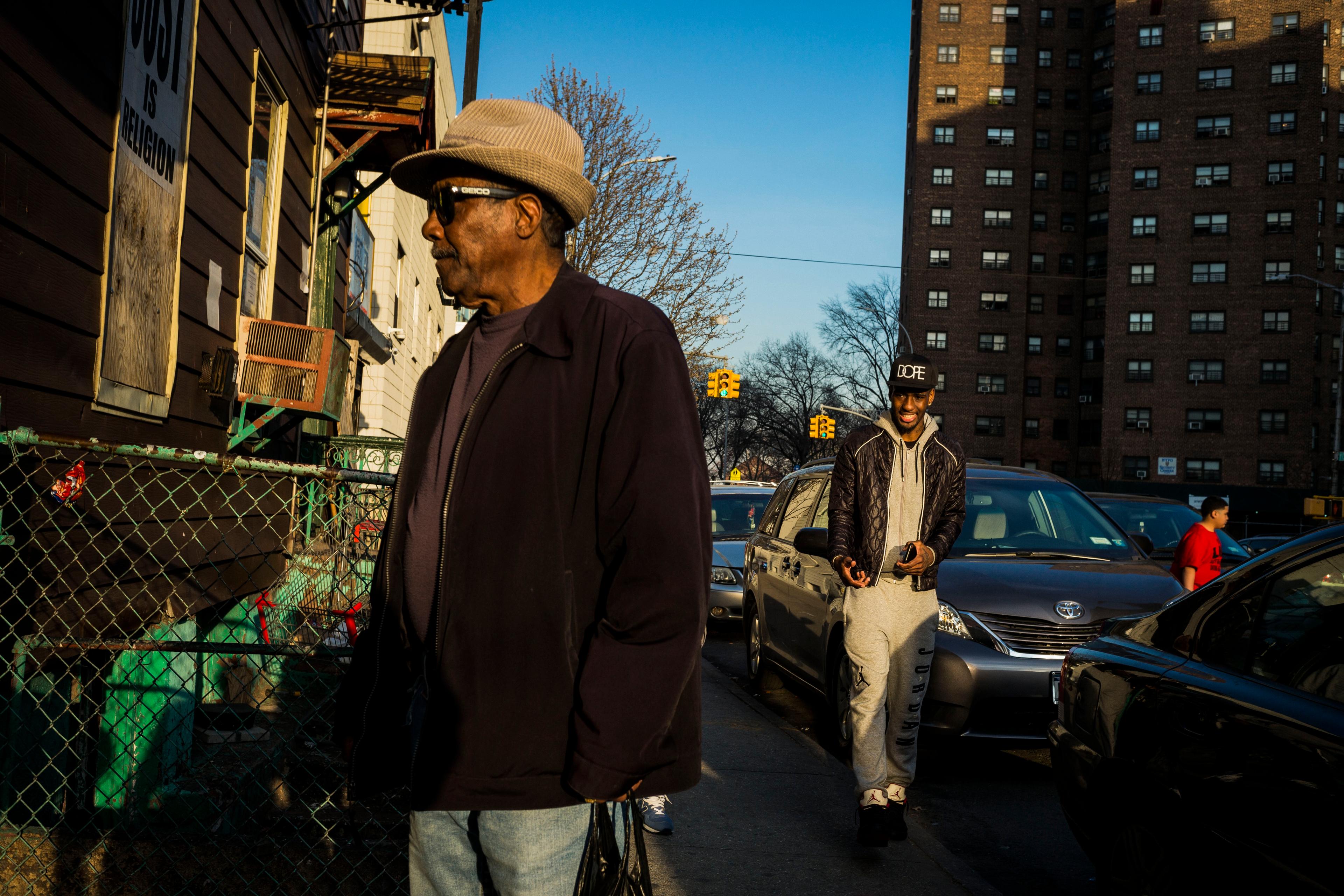Photo of a city street with a man in a hat in the foreground and another man walking among cars in the background.