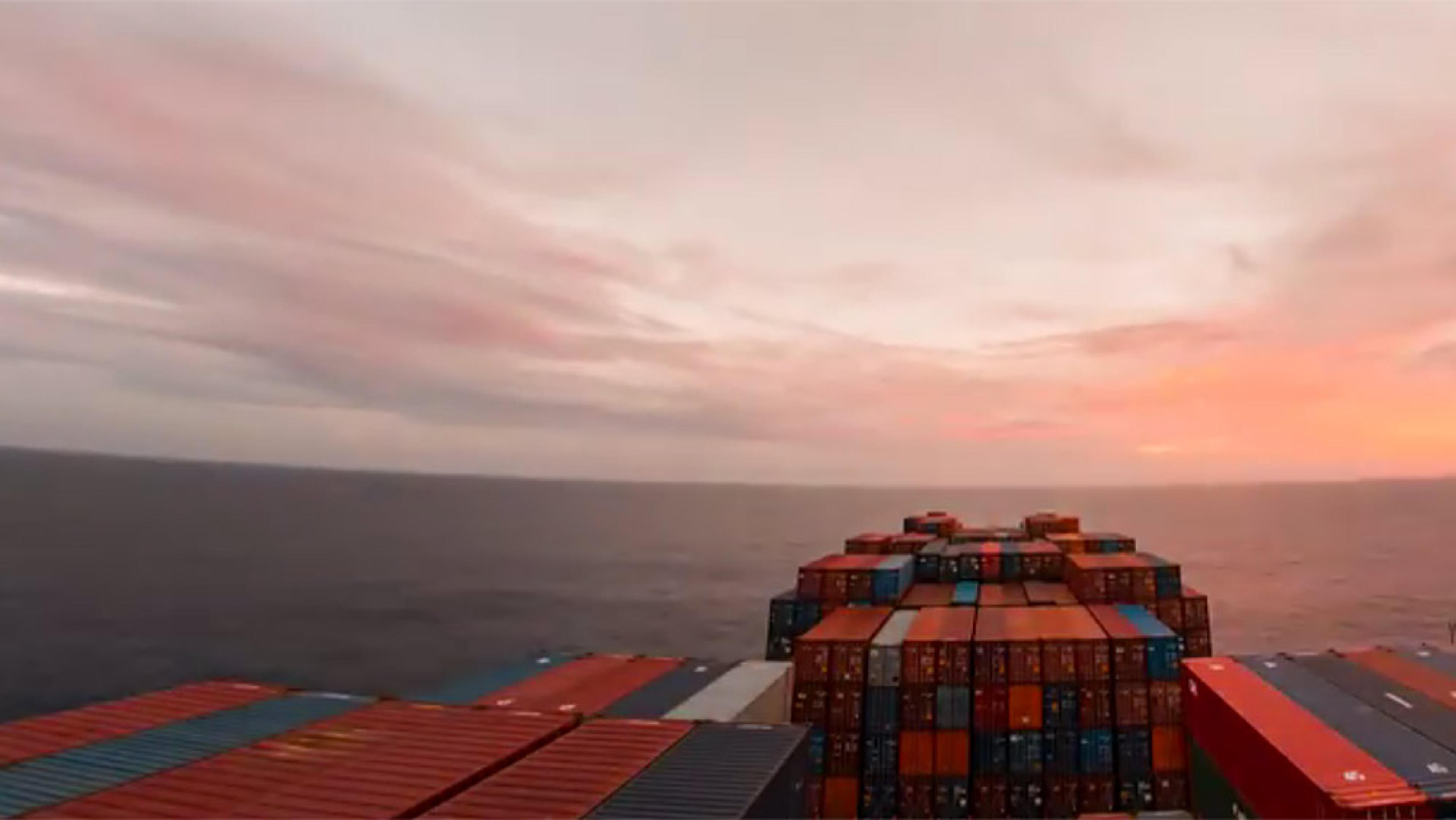 A cargo ship’s deck loaded with colourful shipping containers, sailing on the ocean at sunset with a cloudy sky.