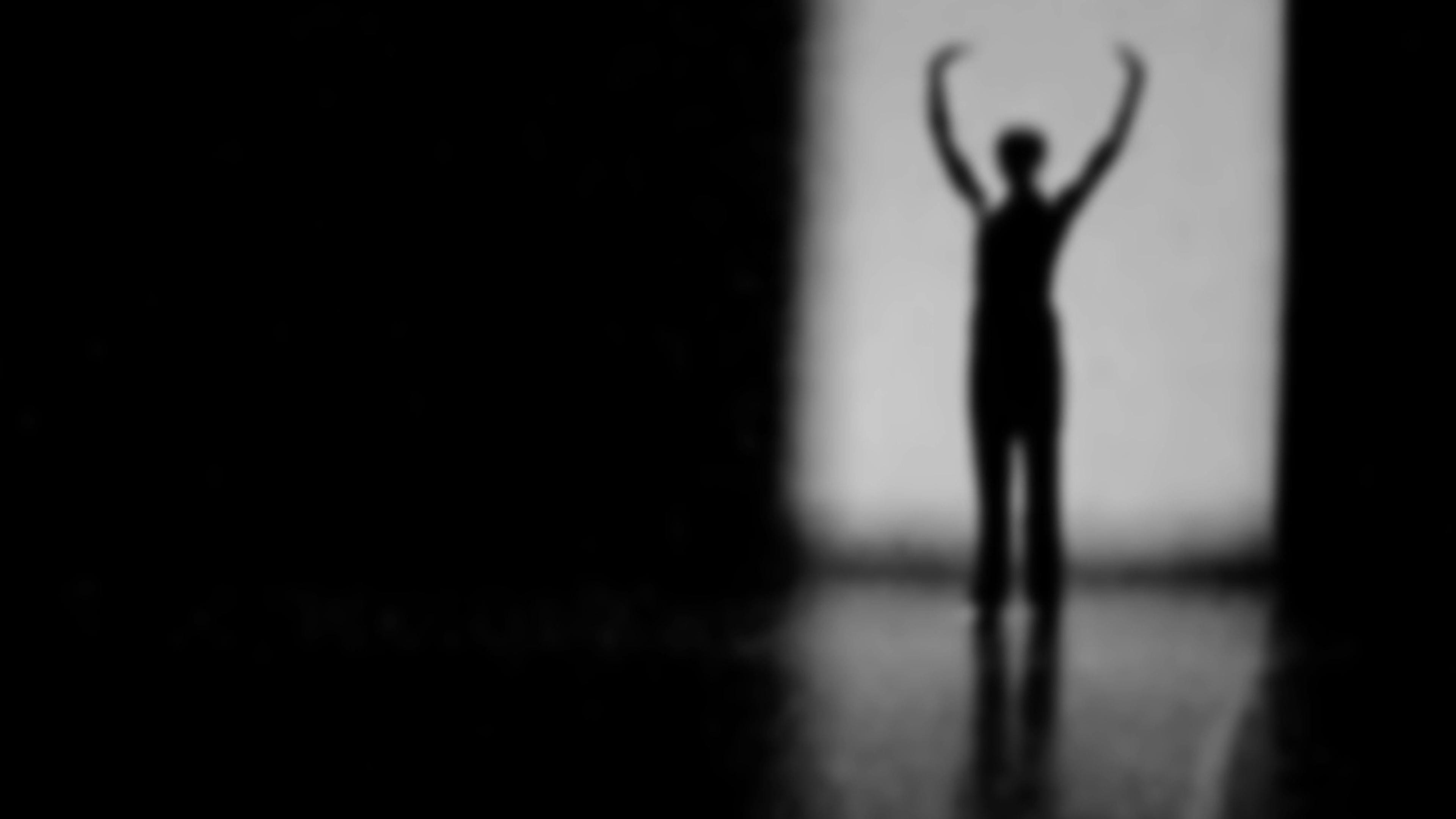 Blurry photo of a person silhouetted with arms raised against a bright rectangular light in a dark setting.