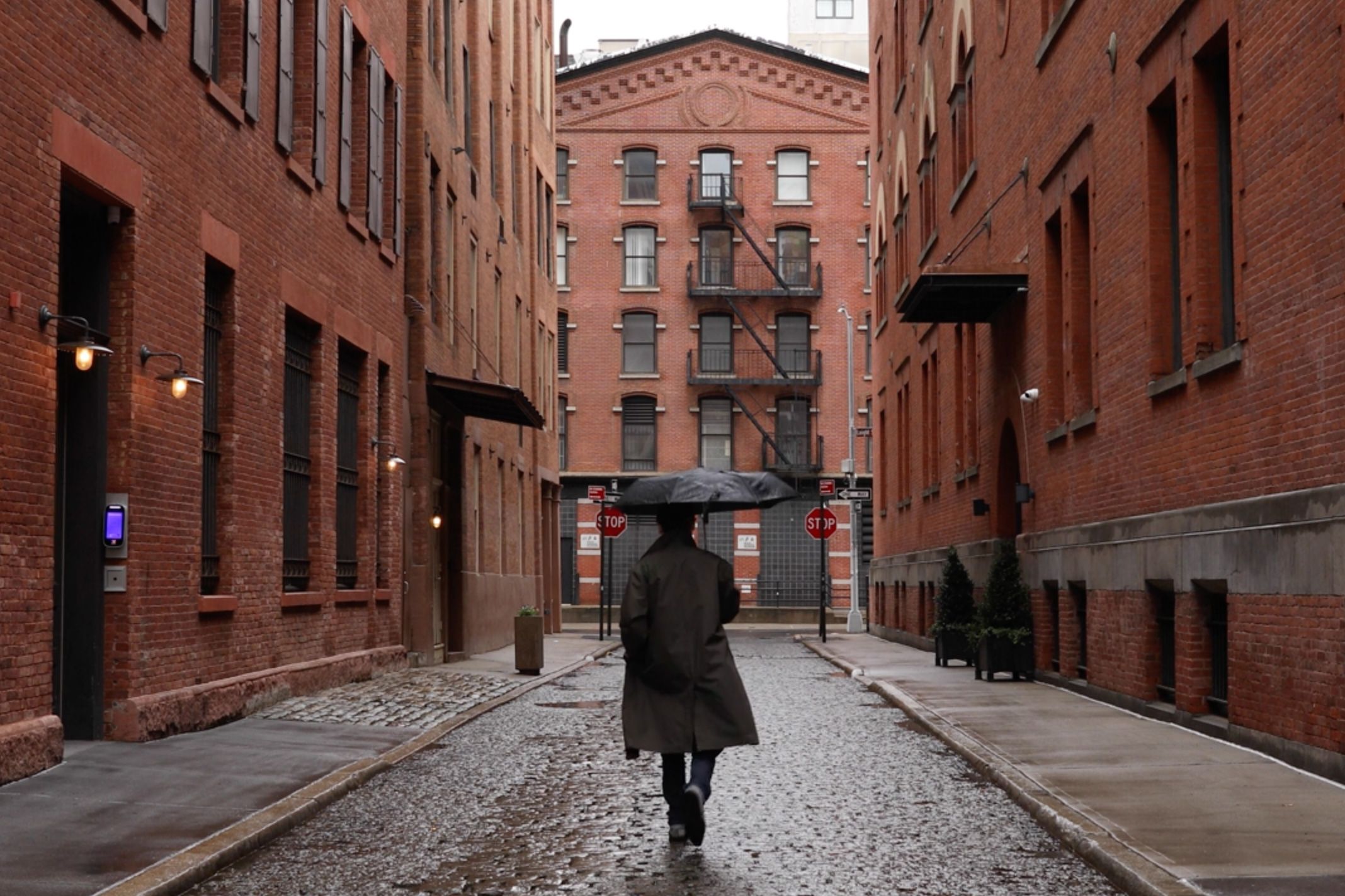 A person with an umbrella walking down a wet brick alley between buildings, with stop signs in the distance.