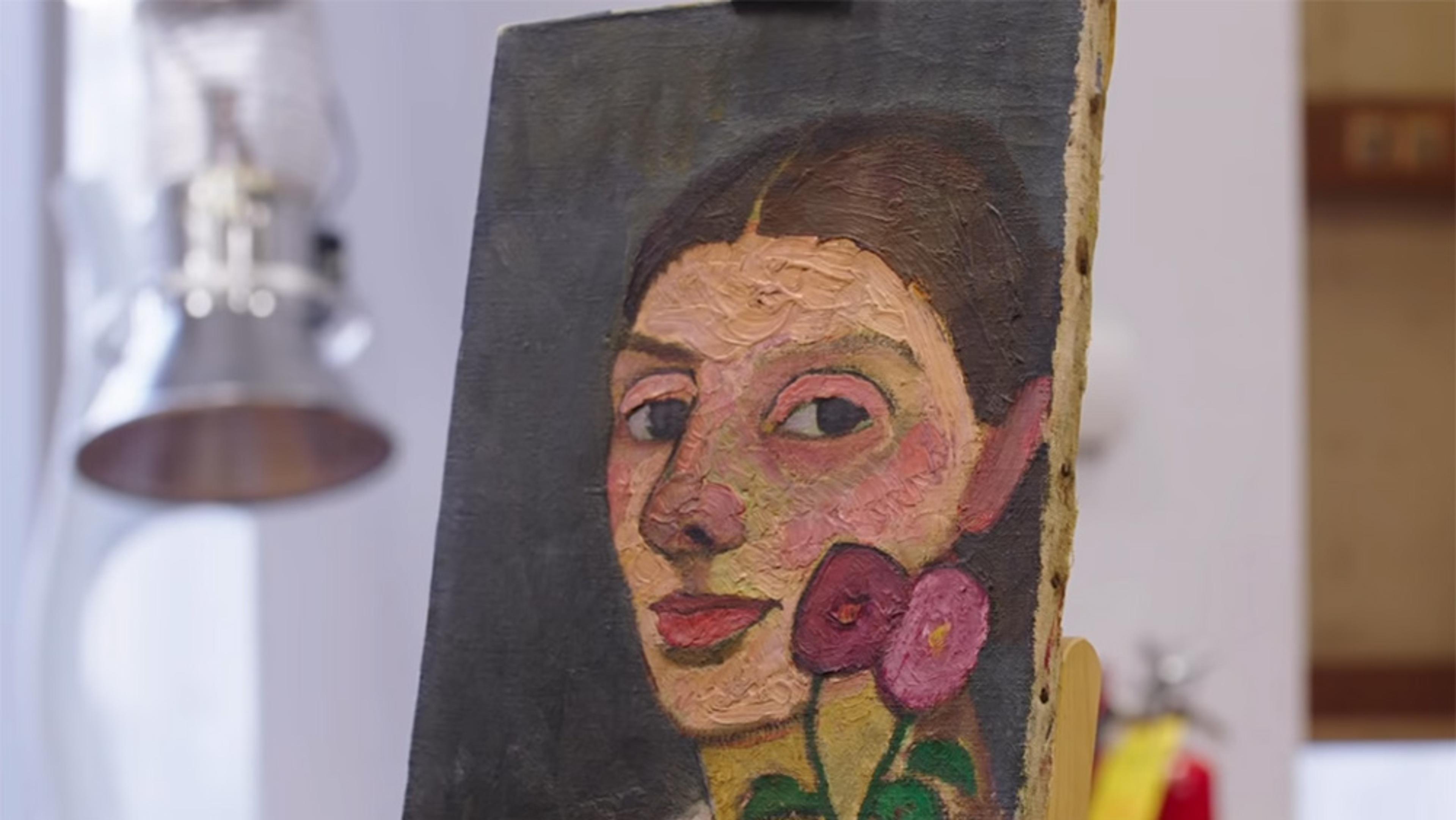 A painting of a woman with two pink flowers is propped up on an easel in a well-lit room.