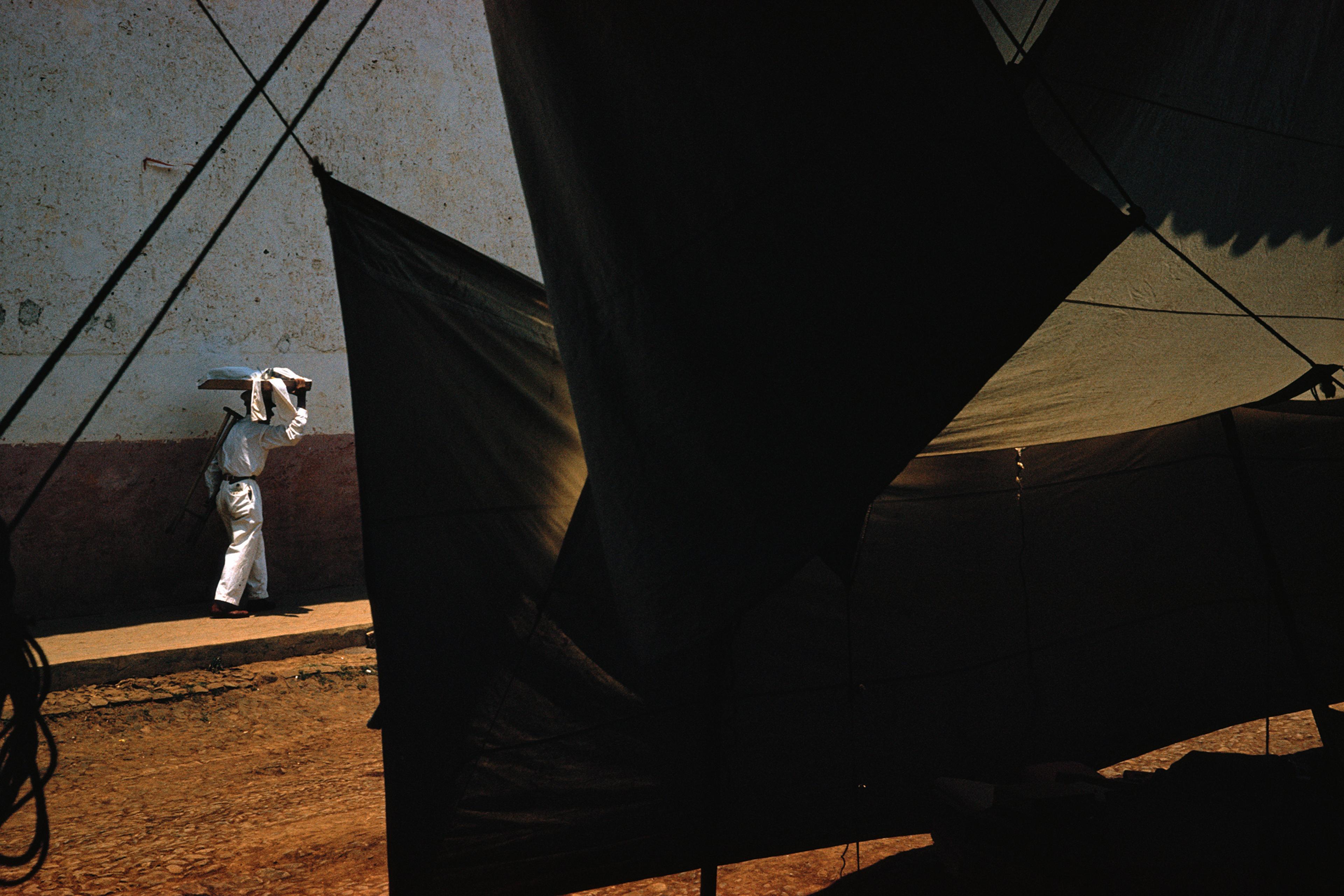 A person in white carrying a load on their head, walking past a dark tent with dramatic light and shadows.