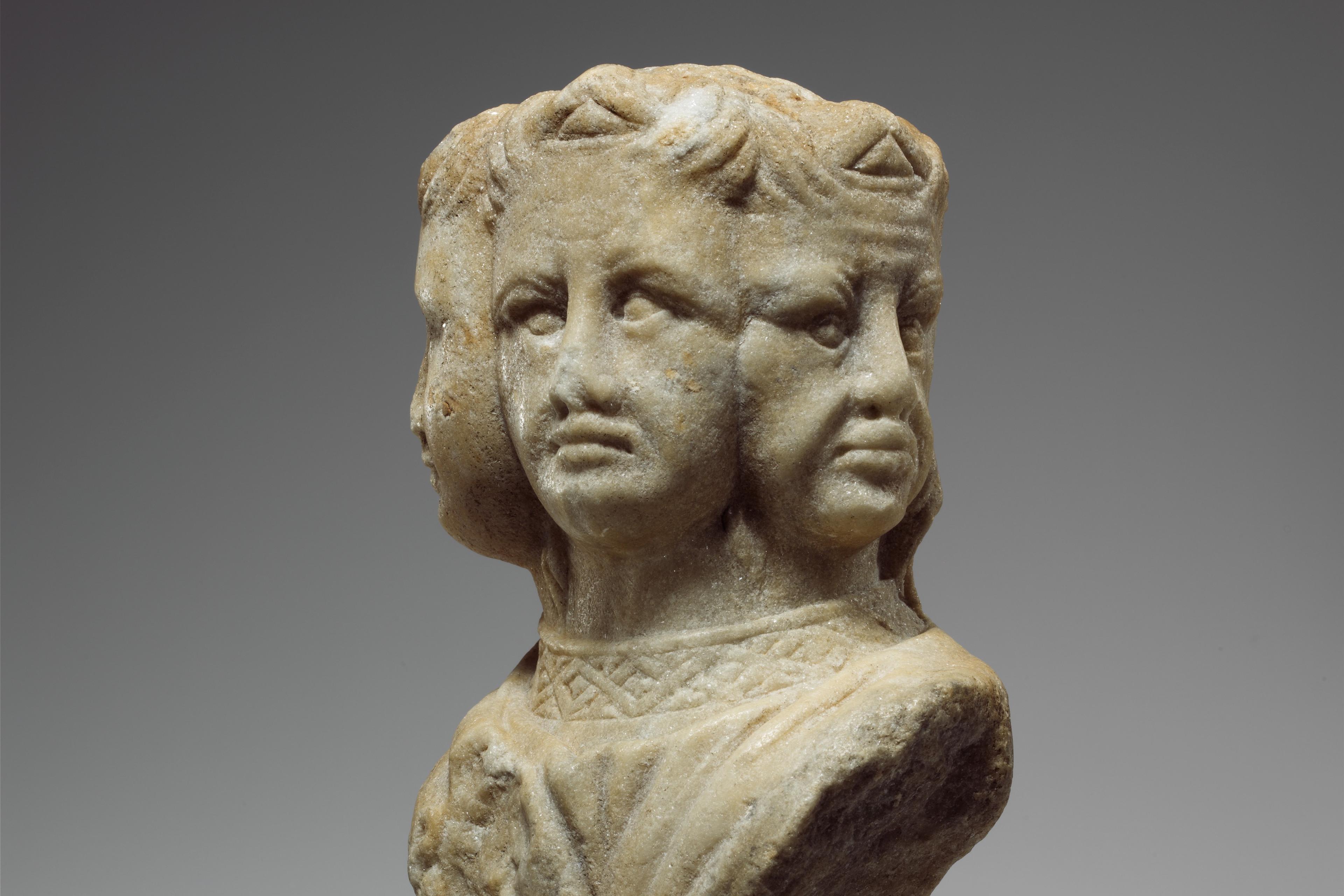 Ancient stone sculpture of three faces on a shared neck. The figure is weathered and detailed, set against a plain grey background.