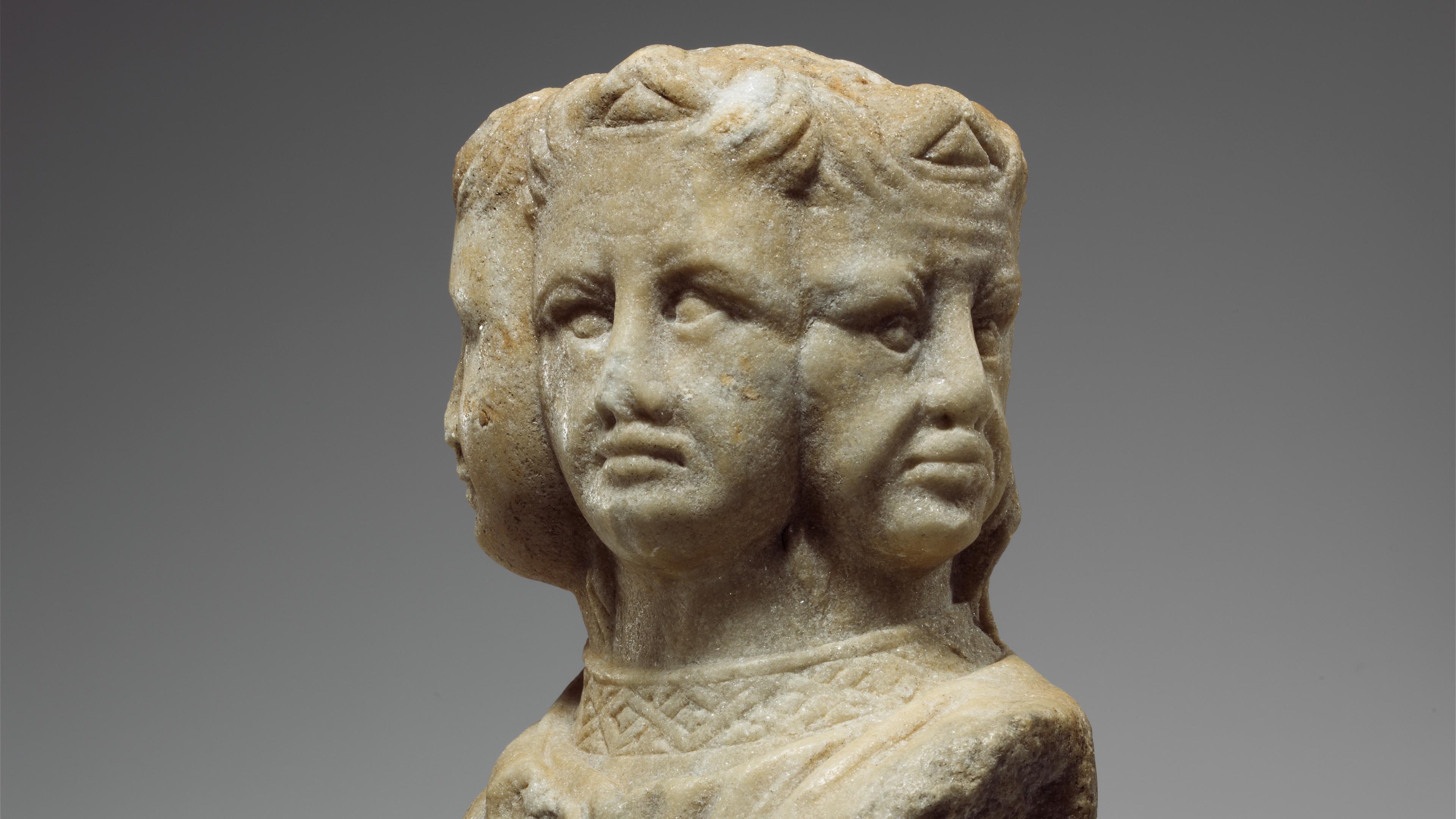 Ancient stone sculpture of three faces on a shared neck. The figure is weathered and detailed, set against a plain grey background.