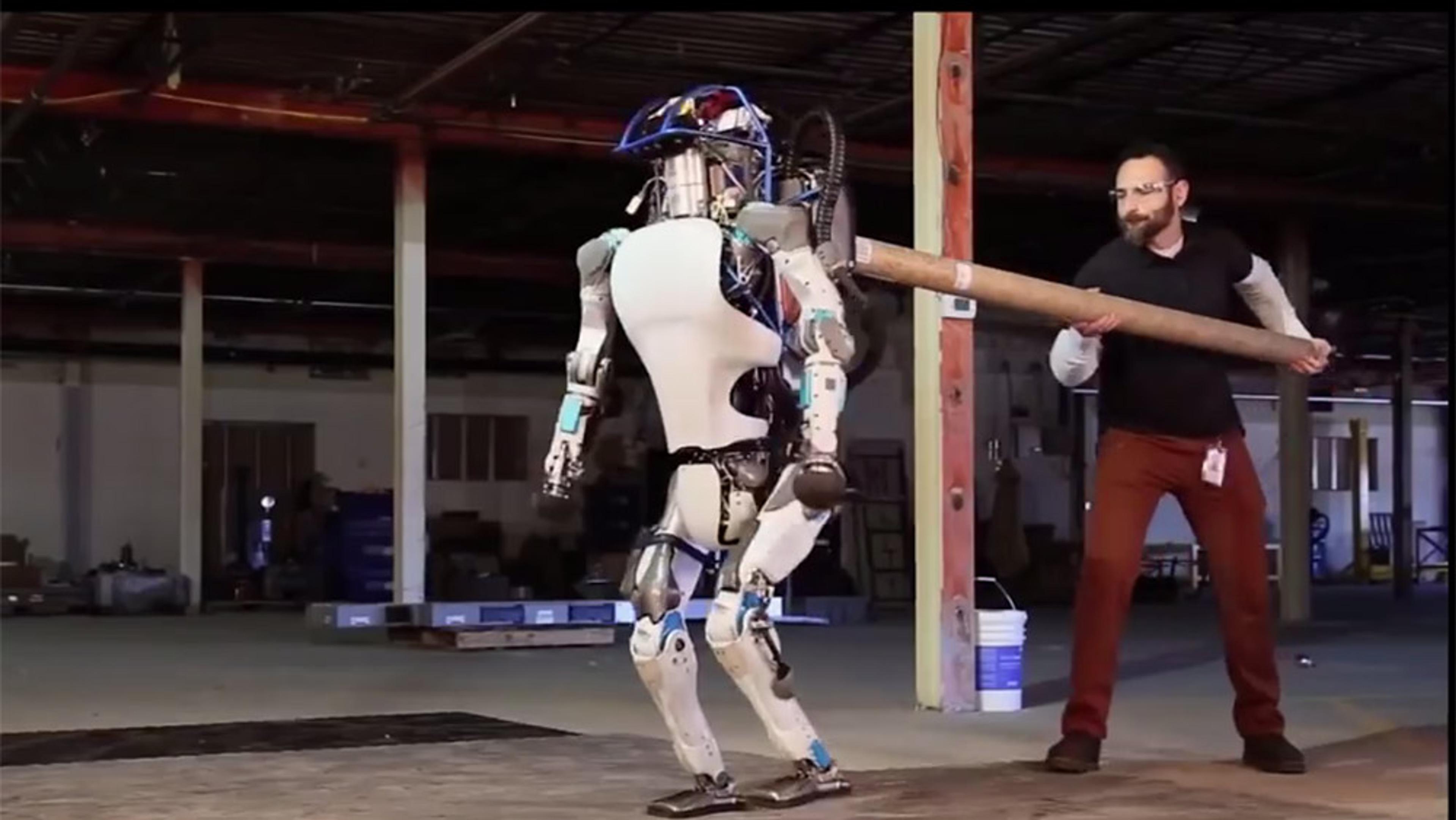 A man testing a humanoid robot’s balance using a pole in an industrial warehouse setting.