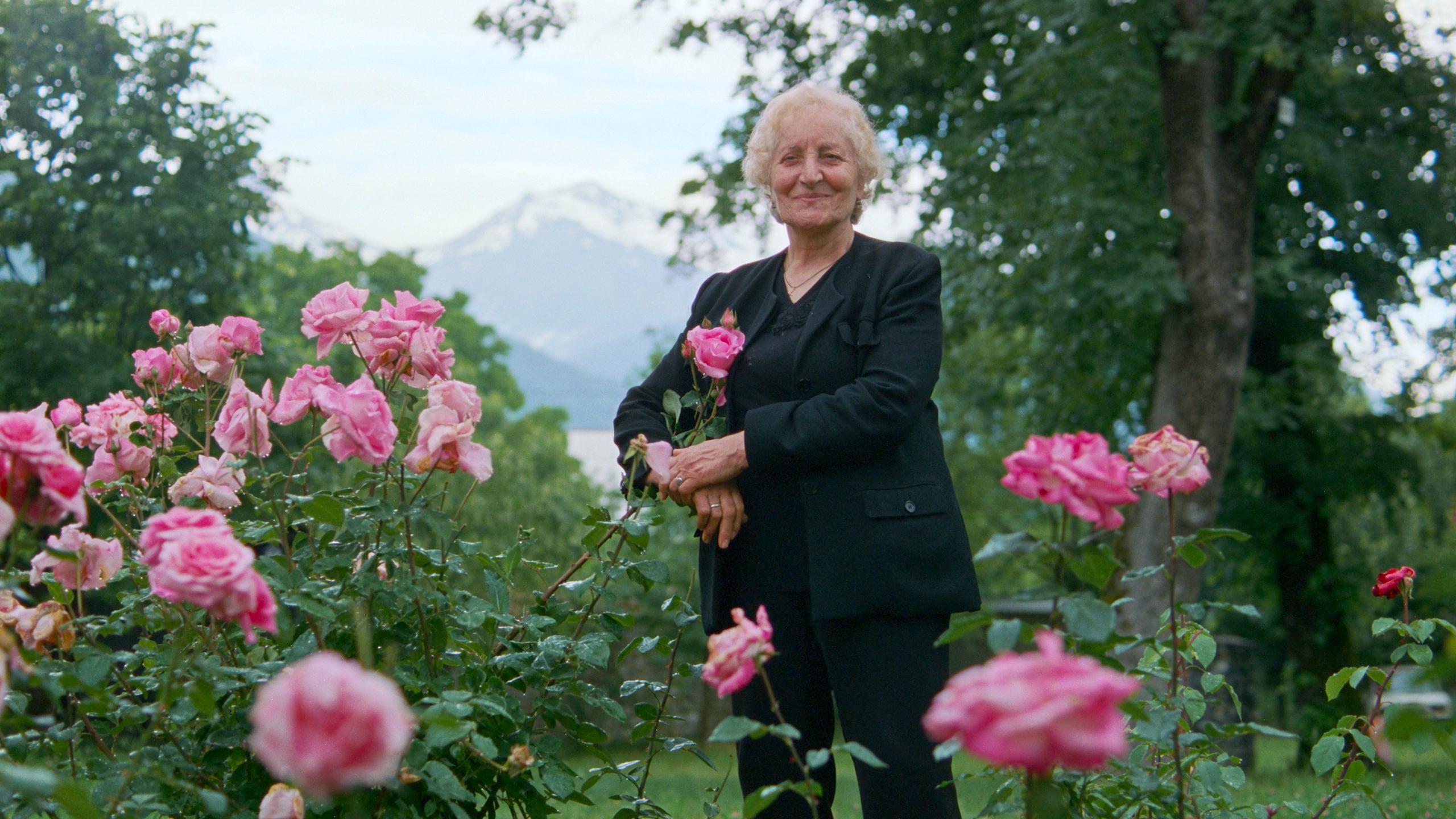 An elderly woman in black standing in a garden of pink roses with snow-capped mountains in the background.