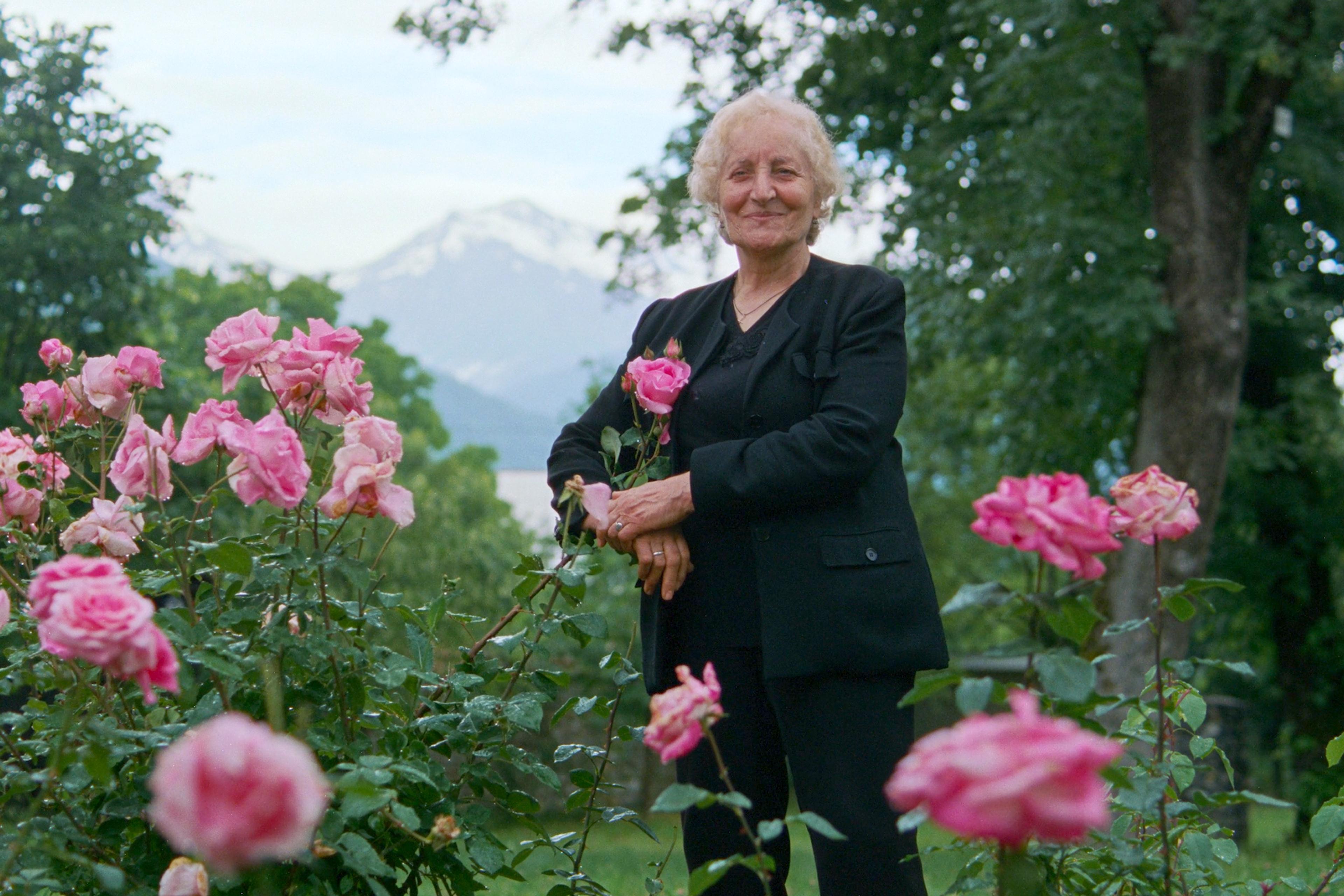 An elderly woman in black standing in a garden of pink roses with snow-capped mountains in the background.
