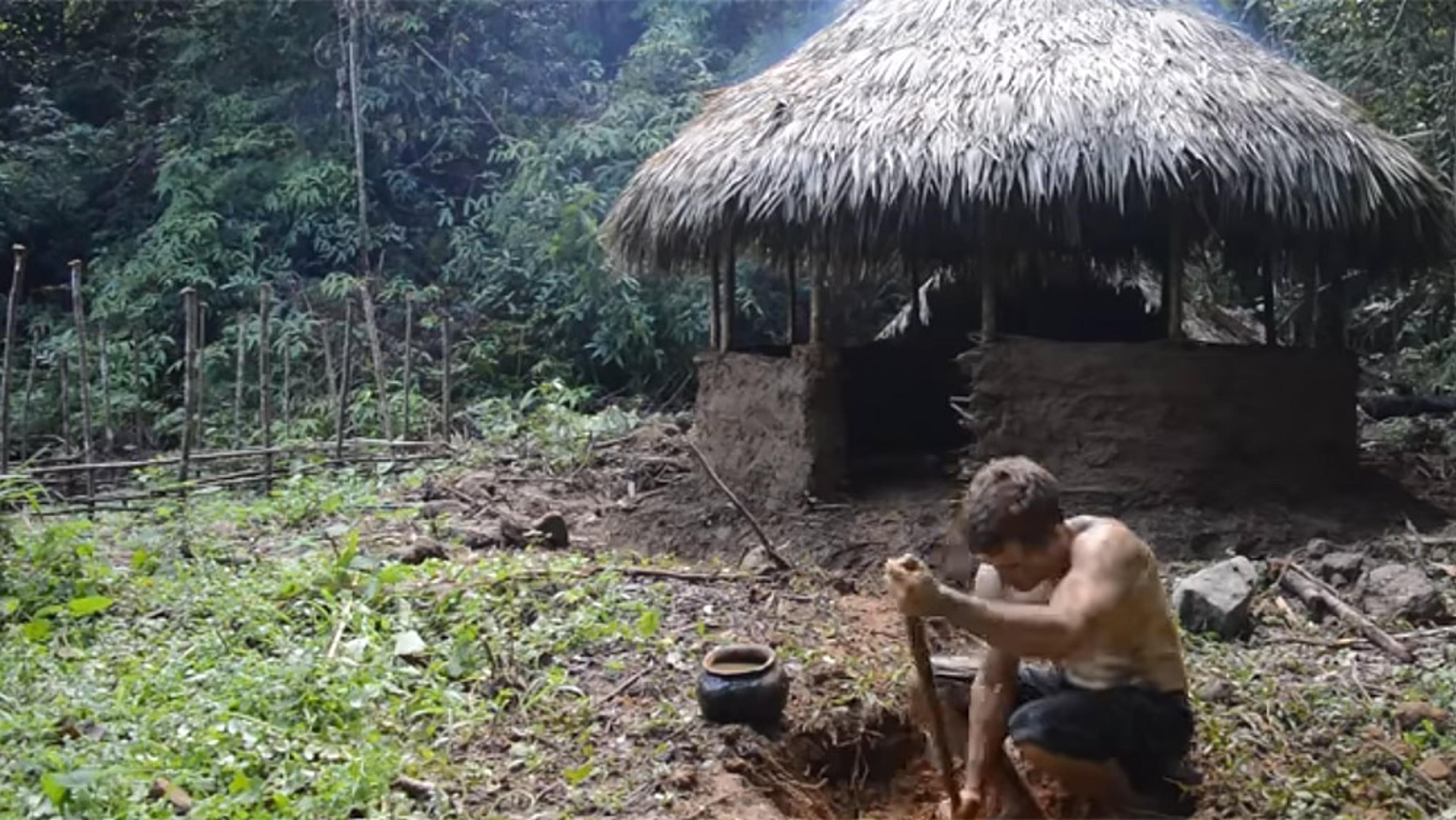 A thatched-roof hut in a jungle, with a man digging in the foreground and a cooking pot nearby.