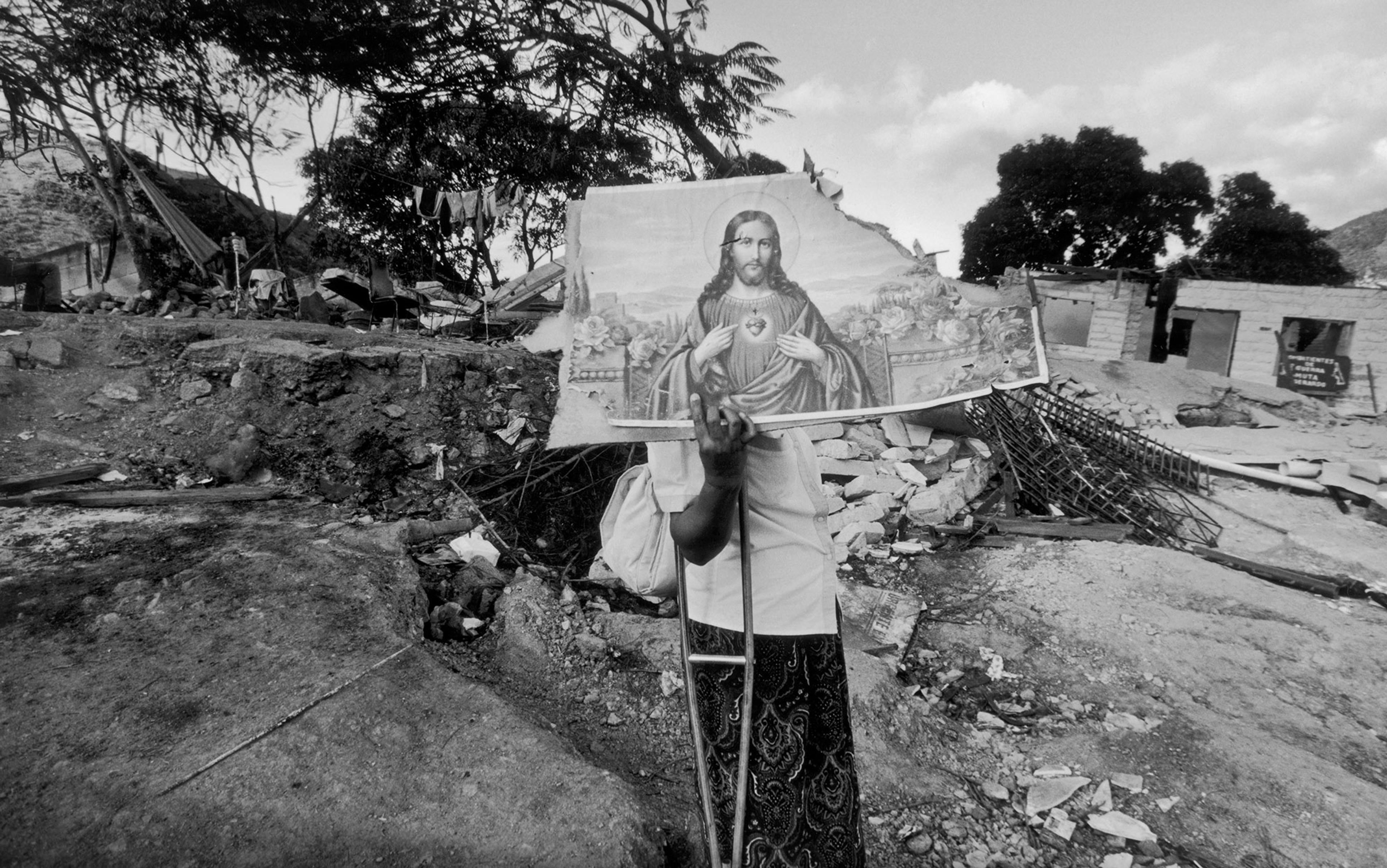 Person holding a painting of Jesus Christ amid a rubble-strewn landscape with damaged buildings and trees in the background.