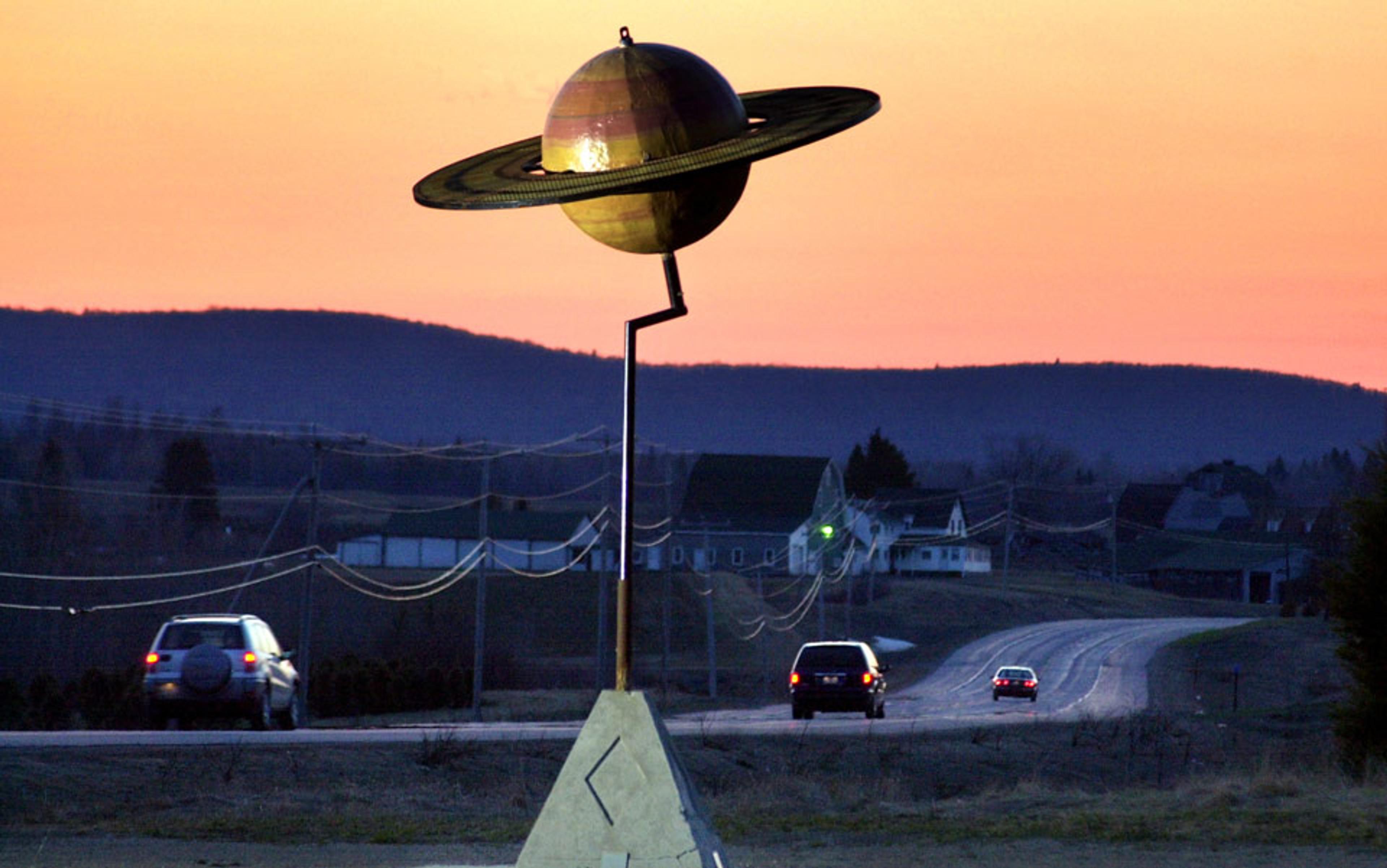 A roadside sculpture of Saturn at sunset, with a curving road and cars in the background and silhouetted hills.