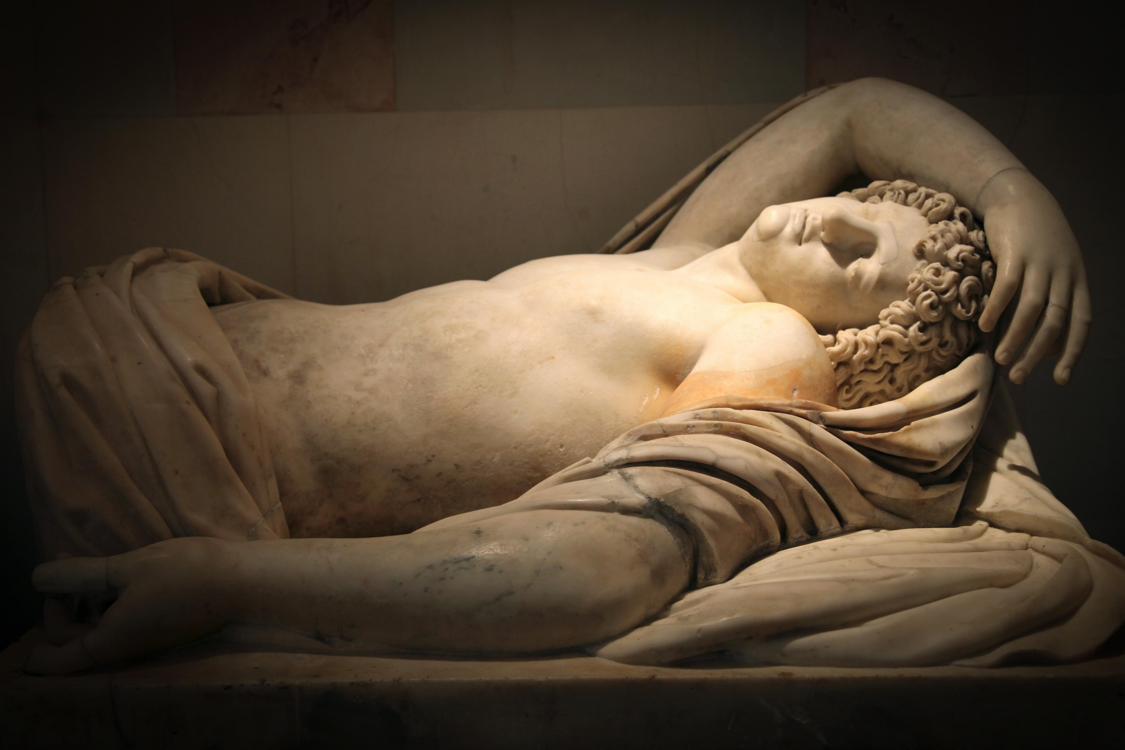 Marble statue of a sleeping man with curly hair lying on fabric, arm draped over his head.