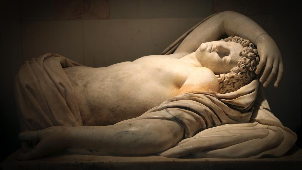 Marble statue of a sleeping man with curly hair lying on fabric, arm draped over his head.