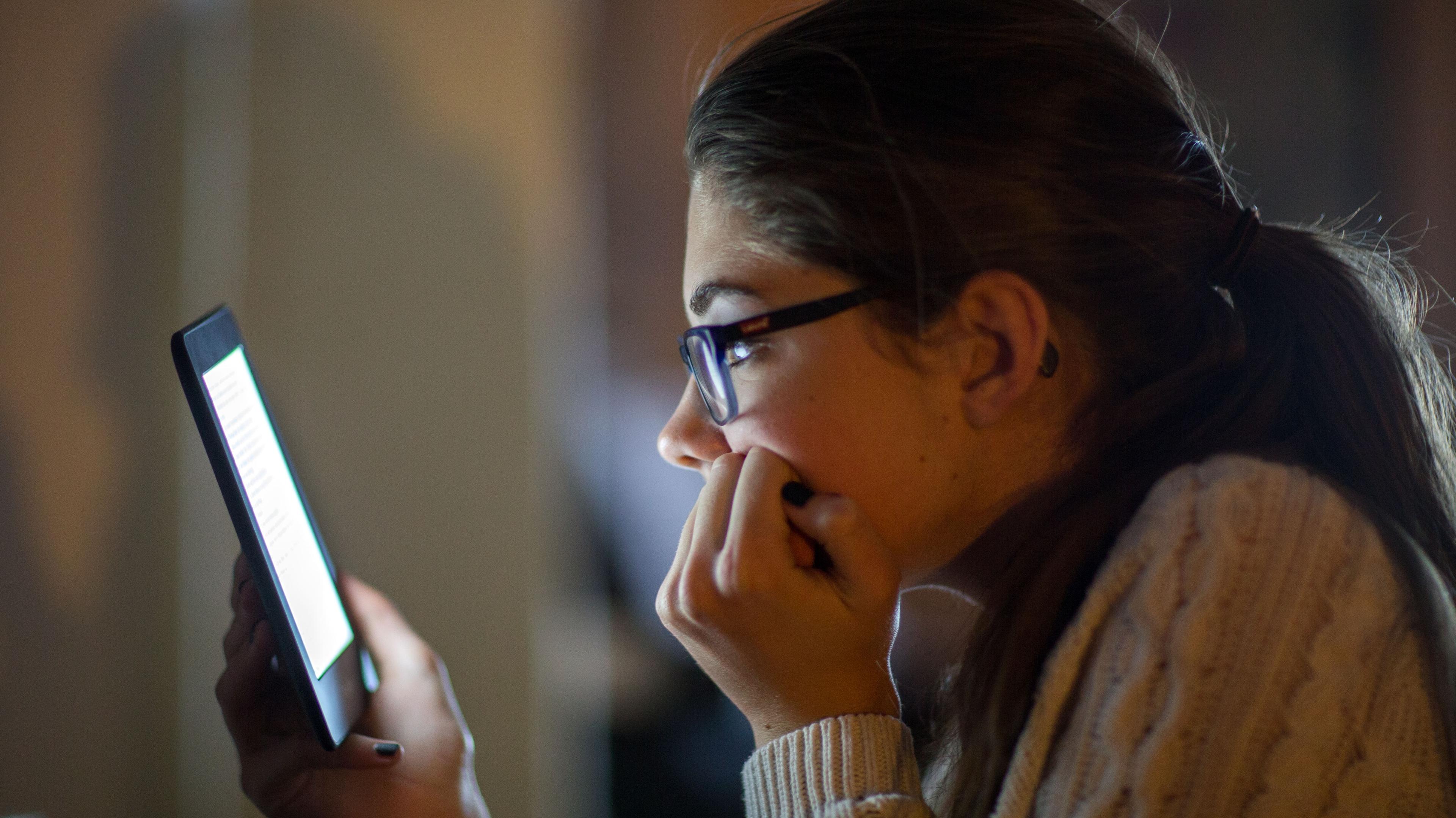 Photo of a person reading on a tablet, facing left, wearing glasses and a jumper in a dimly lit environment.