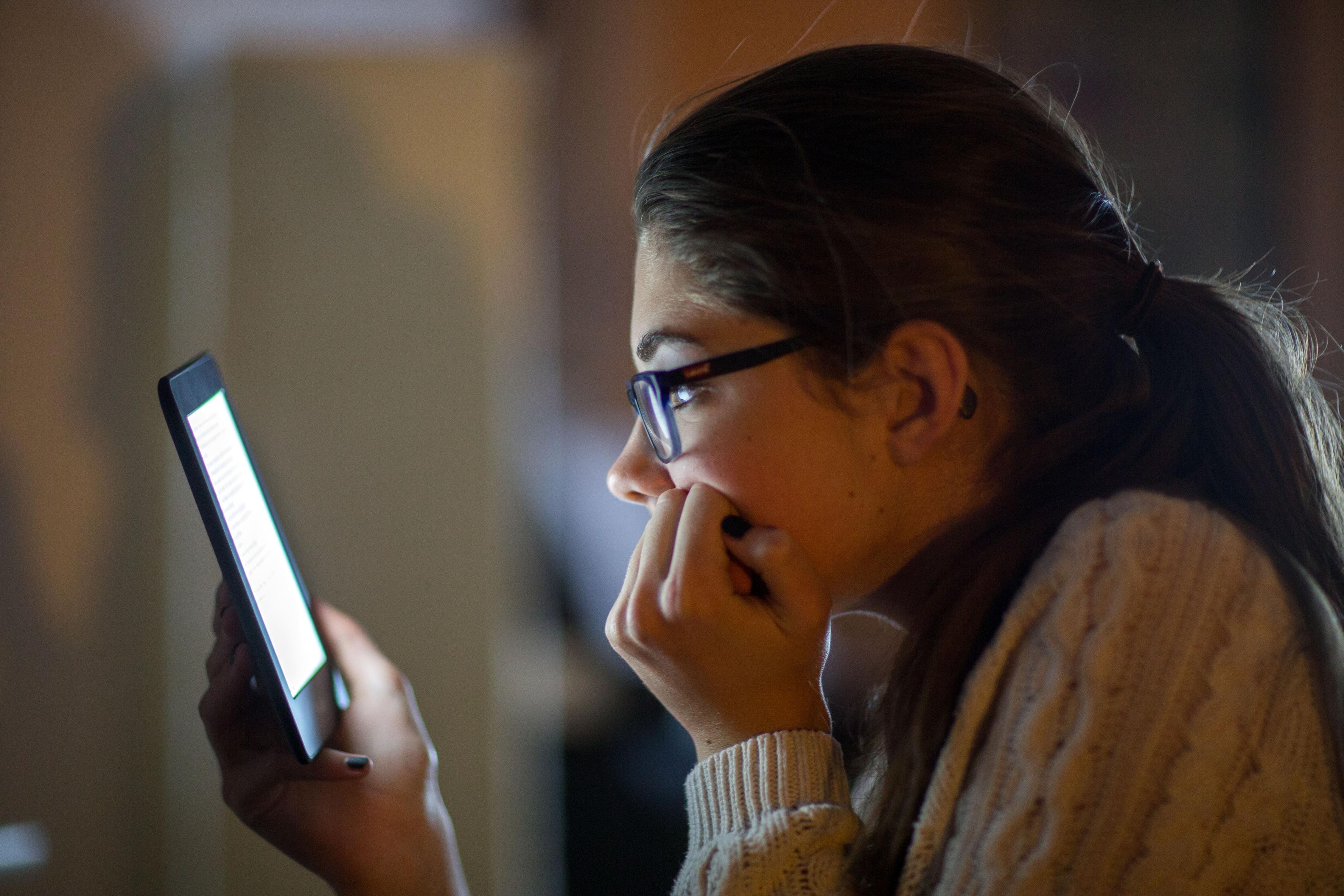 Photo of a person reading on a tablet, facing left, wearing glasses and a jumper in a dimly lit environment.