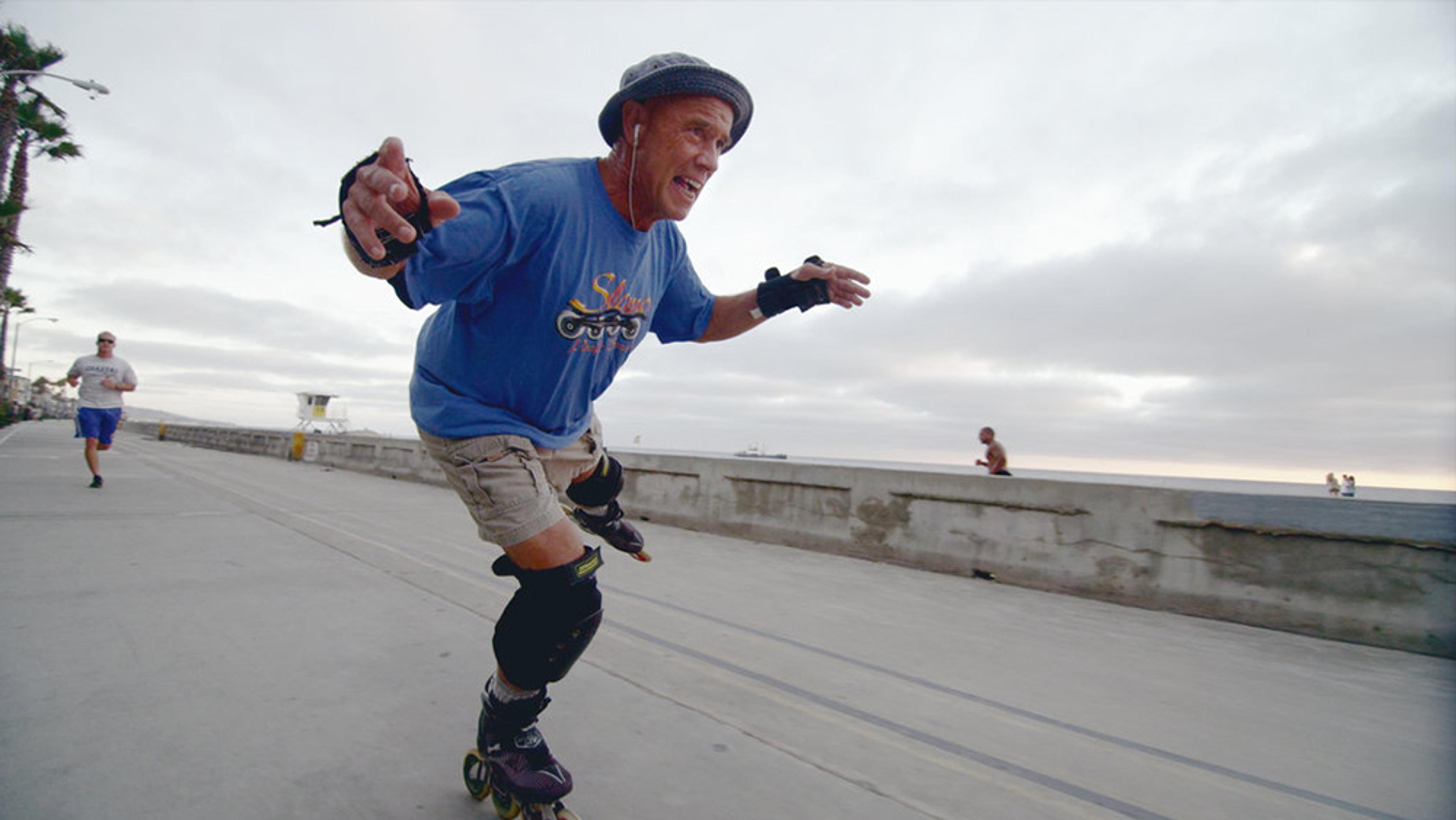An older man rollerblading along a beachfront promenade wearing protective gear and a helmet, with joggers in the background.