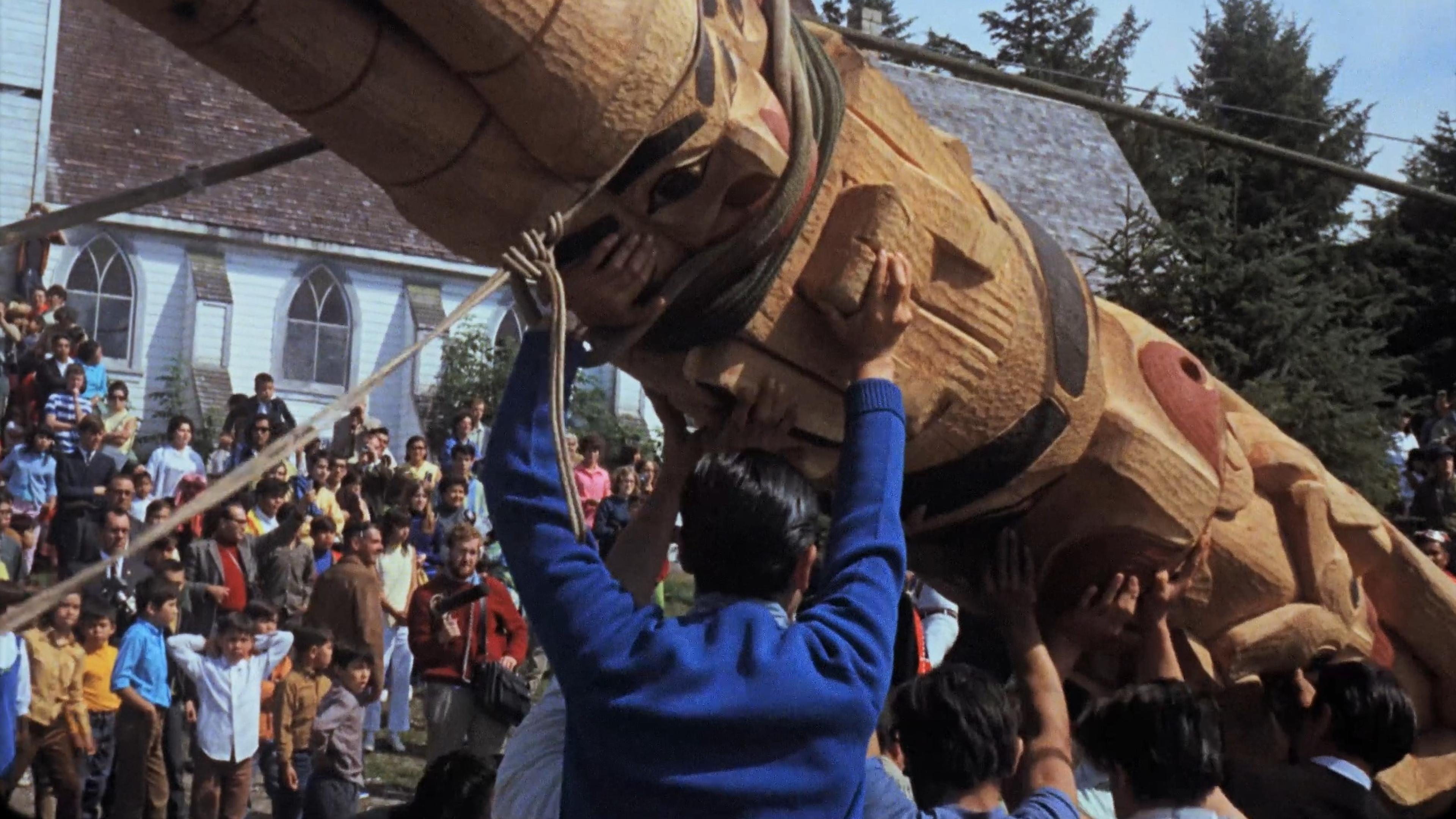 A crowd raising a large totem pole in front of a church with arched windows while children and adults look on.