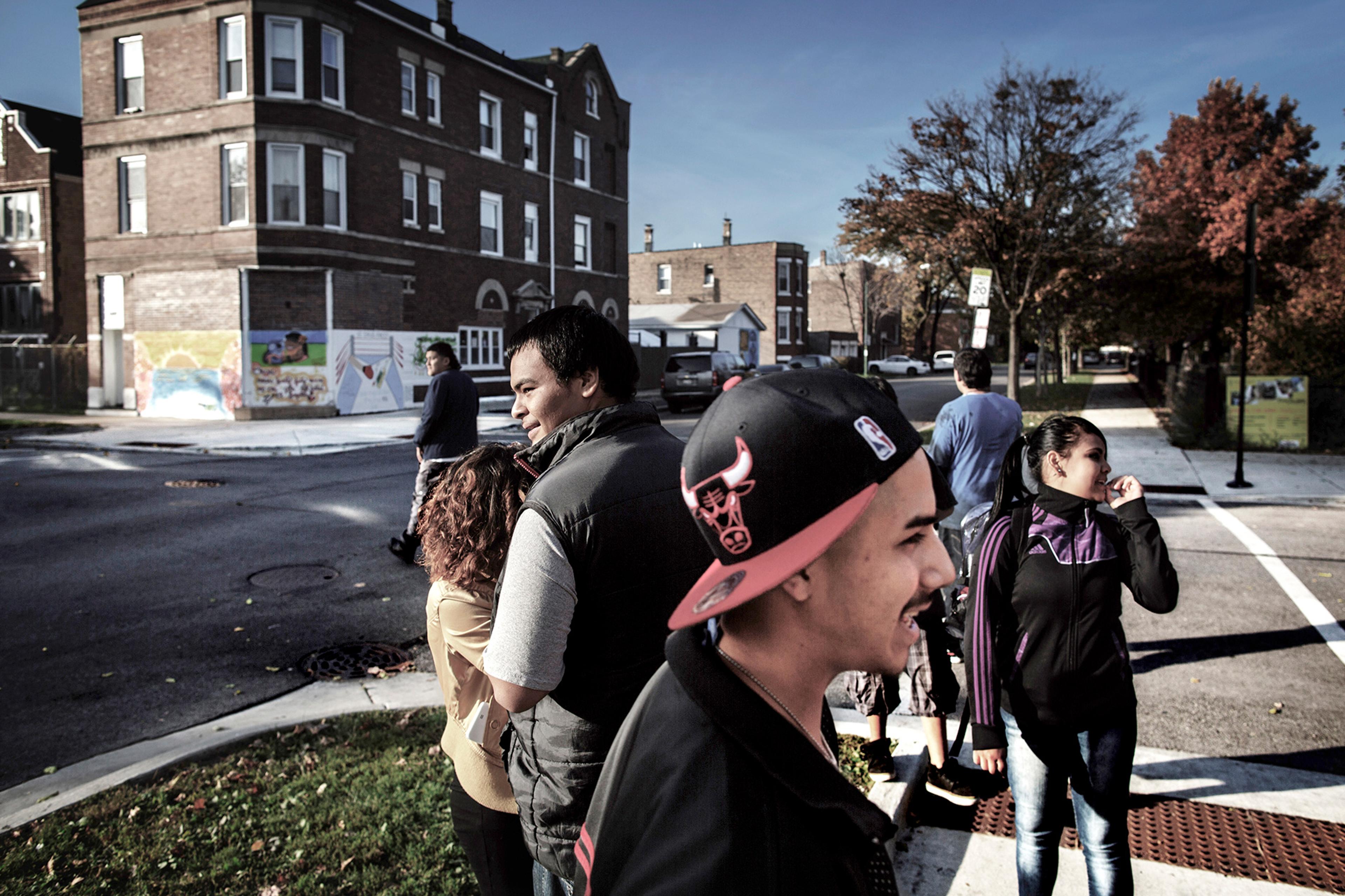 People standing and walking near a street corner with a brick building and a colourful mural in the background on a sunny day.