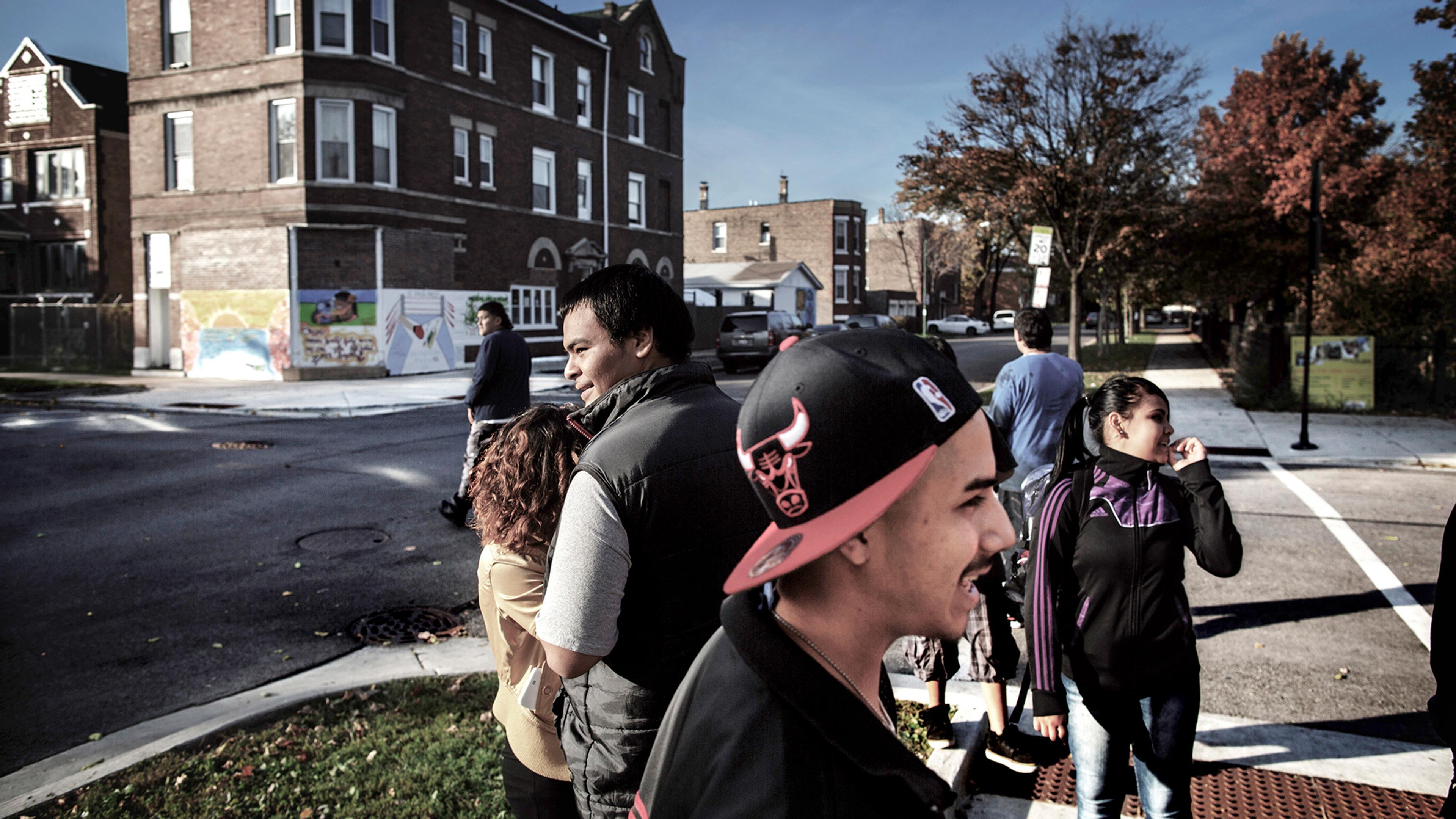 People standing and walking near a street corner with a brick building and a colourful mural in the background on a sunny day.