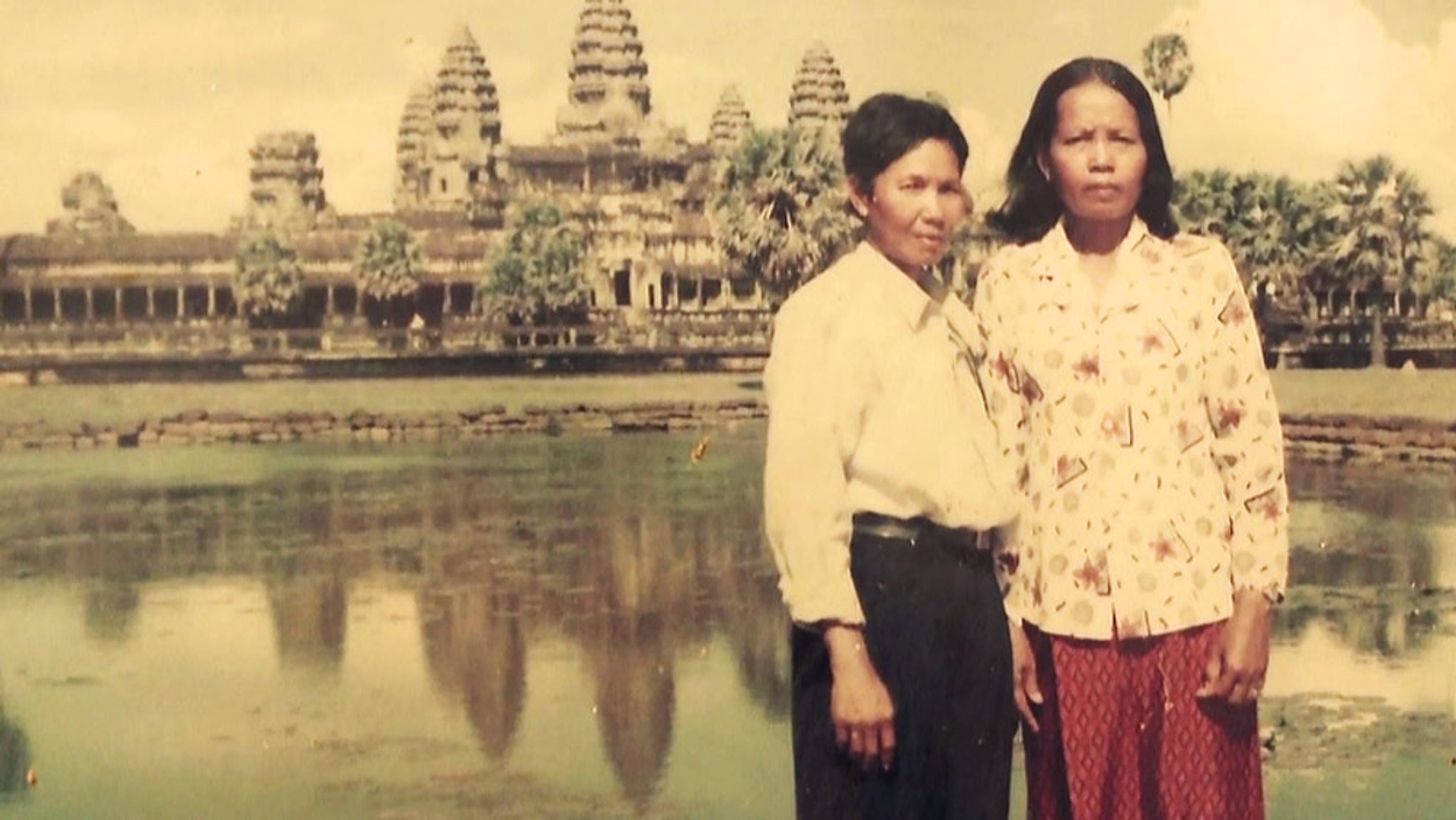 A vintage photo of two Asian women standing in front of Angkor Wat with a reflective pool in the foreground and cloudy skies above.