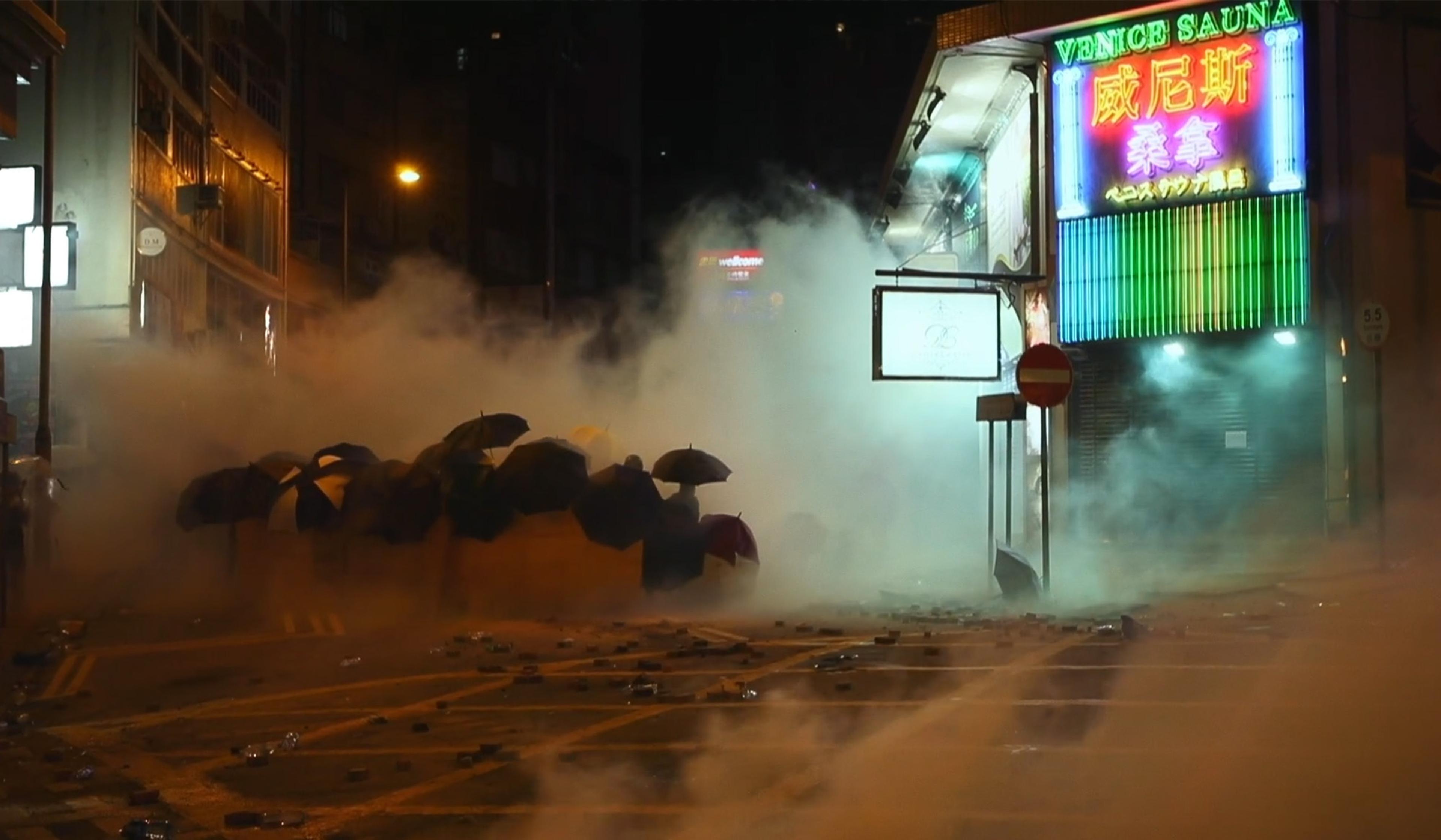 A nighttime street scene including a group of people with umbrellas amid haze and debris. Smoke fills the air near a neon-lit building that says “Venice Sauna”.