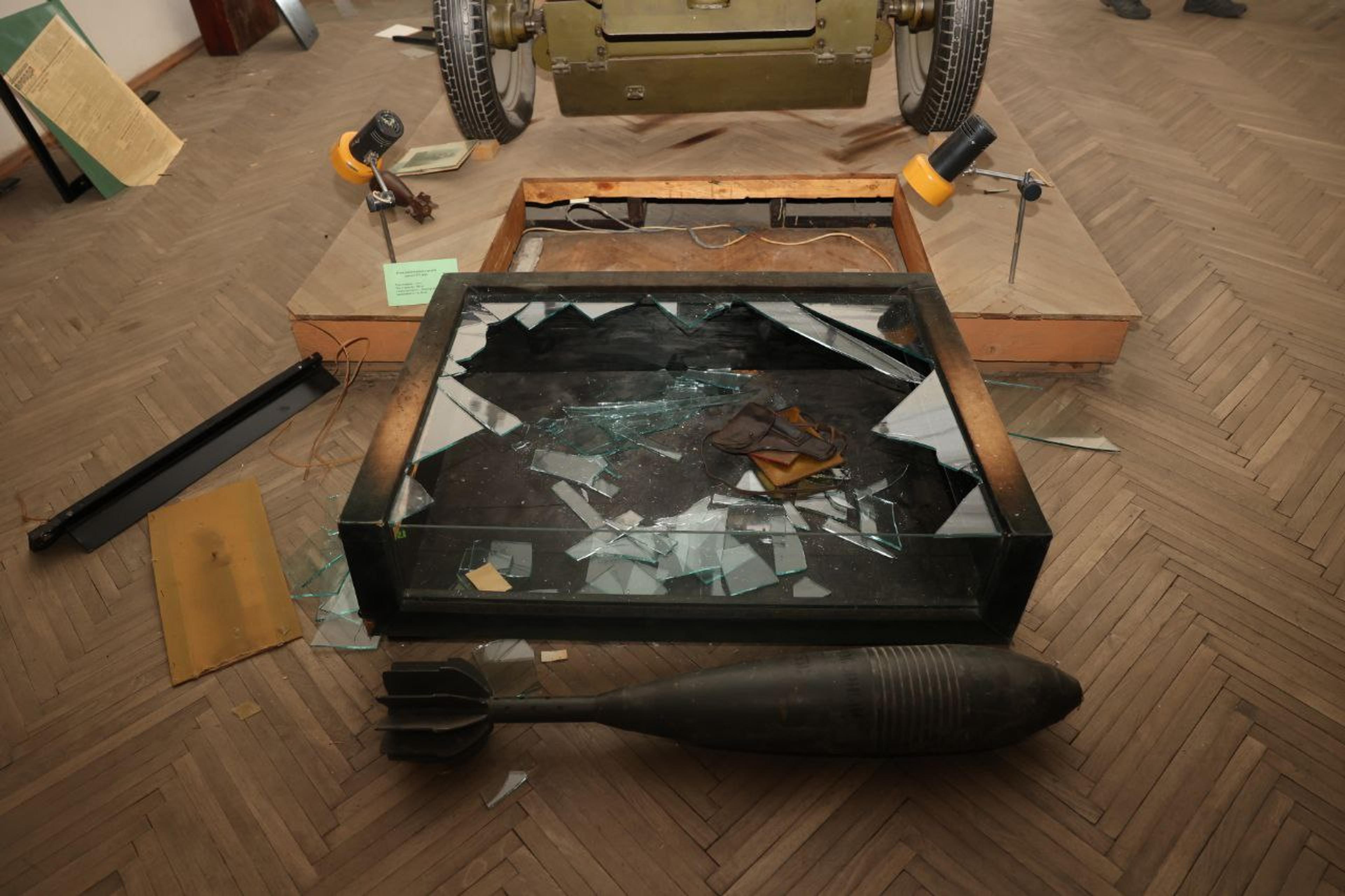 A broken display case with shattered glass, a pair of gloves inside, a bomb beside it and a green military vehicle in the background.