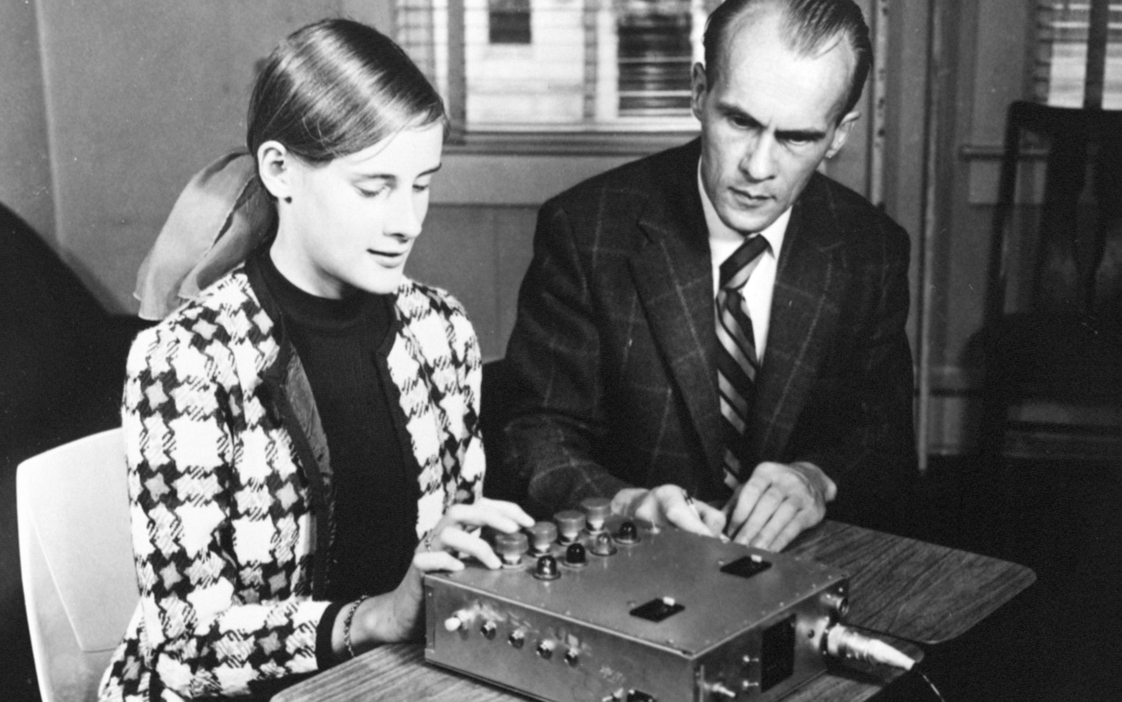 A young woman and a man, both dressed formally, sit at a table with an electronic device in front of them. The woman is engaging with the device, which has several buttons and dials, while the man observes attentively. The setting appears to be an office or classroom. The image is in black and white.