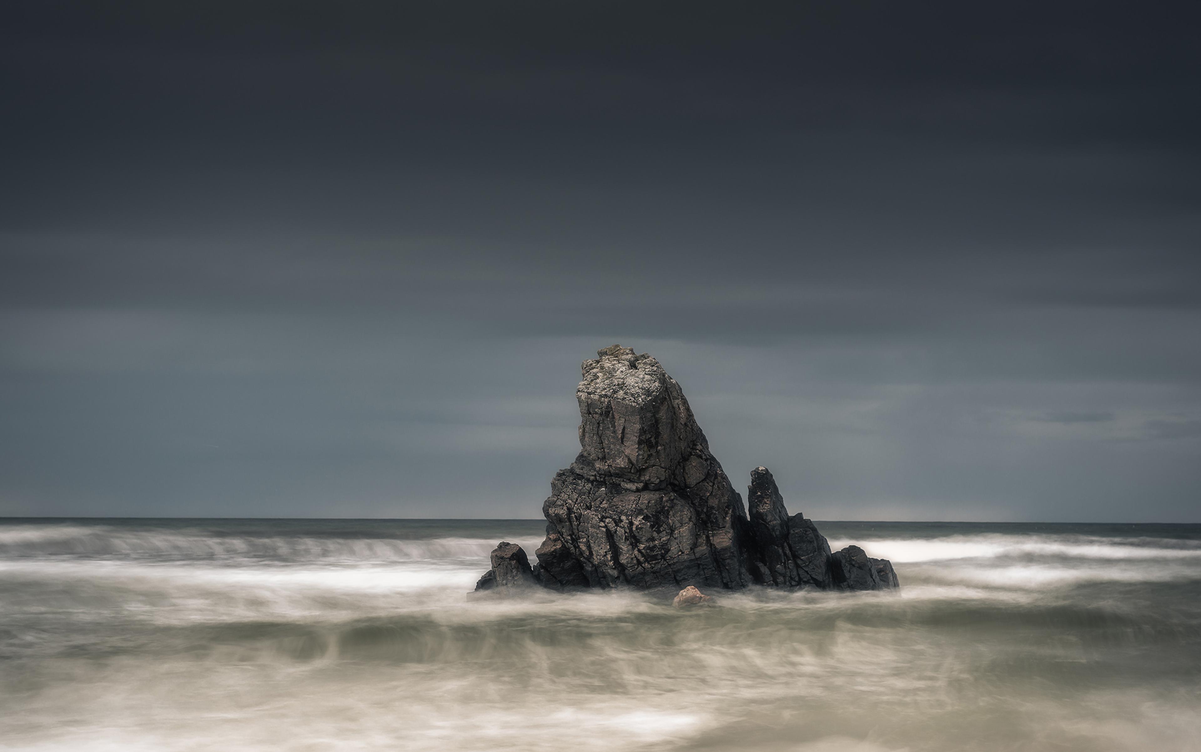 Photo of a dark, rugged rock formation emerging from blurred ocean waves under a cloudy sky.