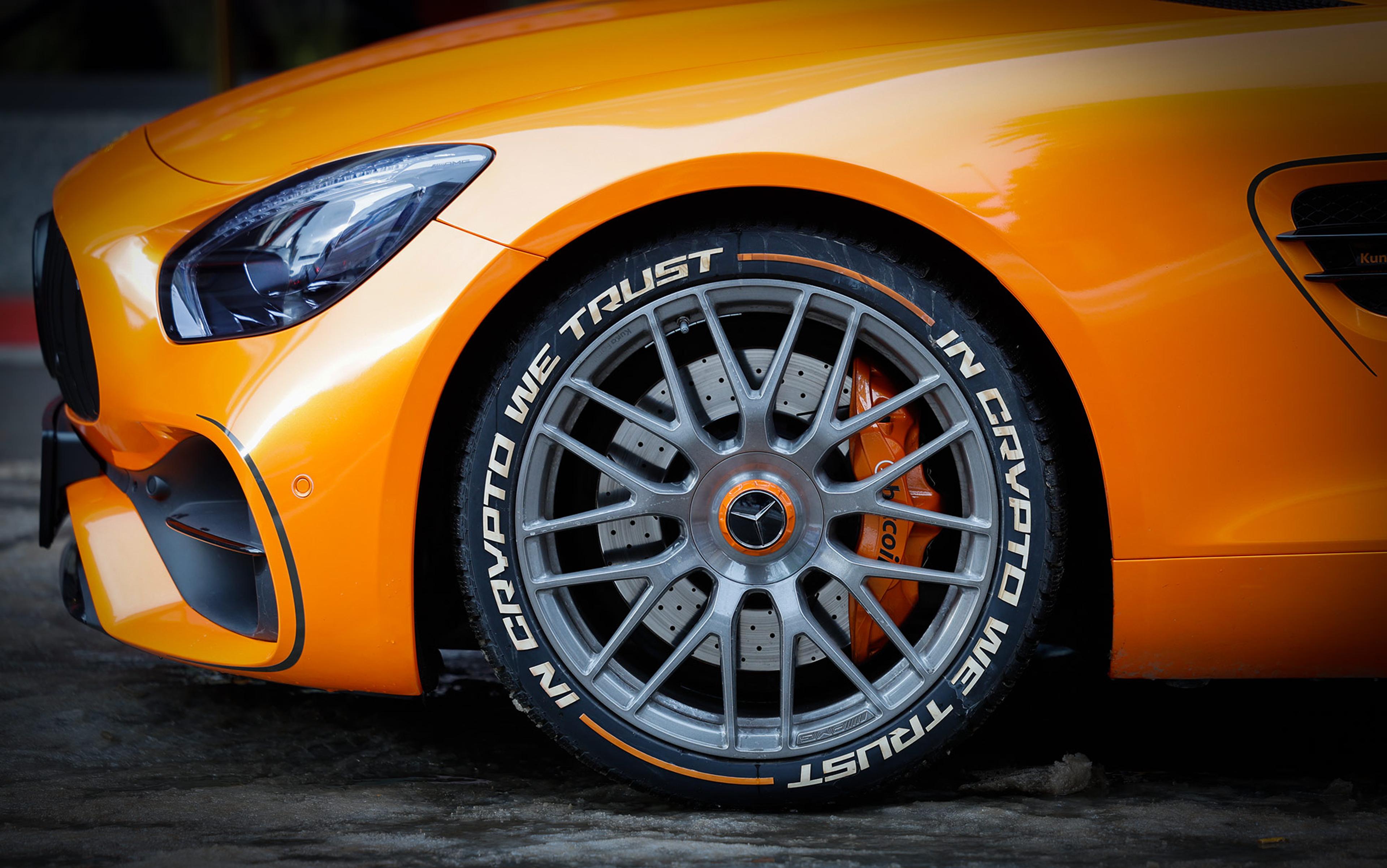Close-up of an orange Mercedes car with the focus on the front tyre, which is inscribed with ‘In Crypto We Trust’