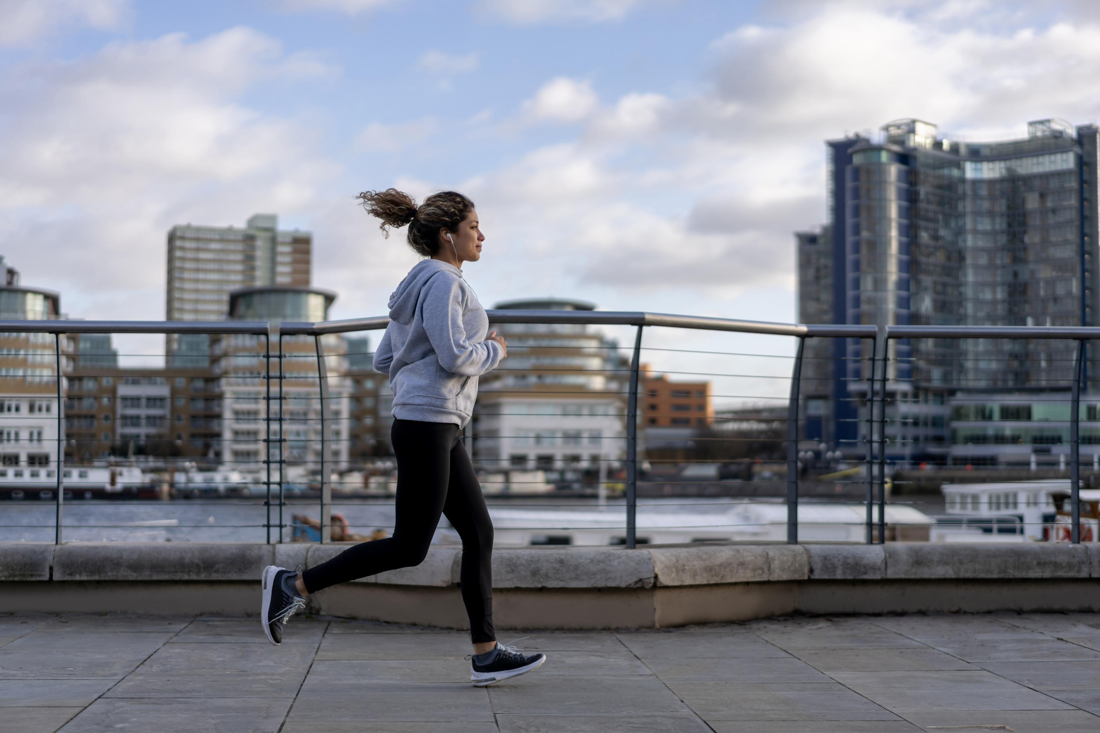 Photo of a woman jogging along a riverside path in an urban area with tall buildings under a partly cloudy sky.