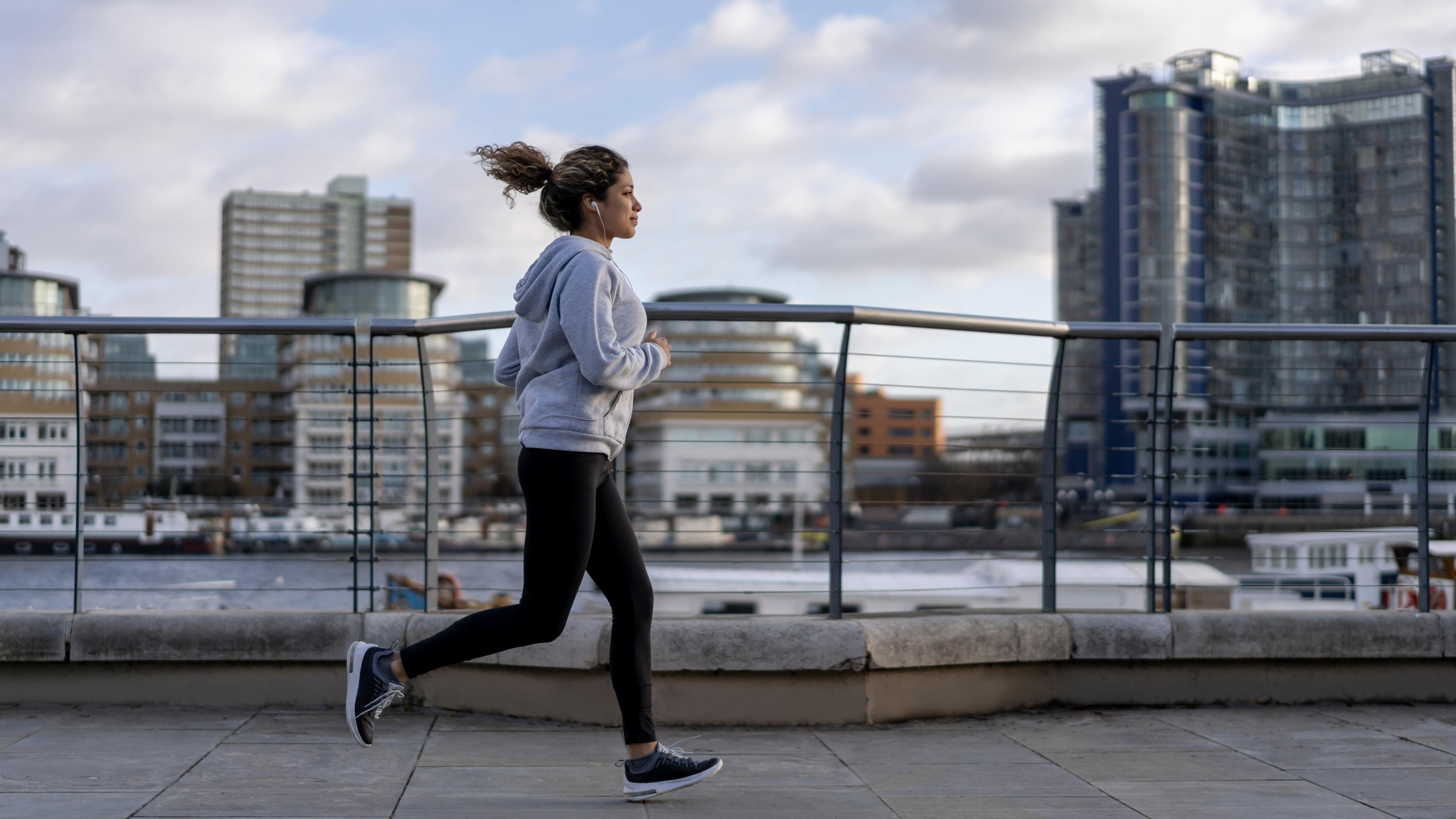 Photo of a woman jogging along a riverside path in an urban area with tall buildings under a partly cloudy sky.